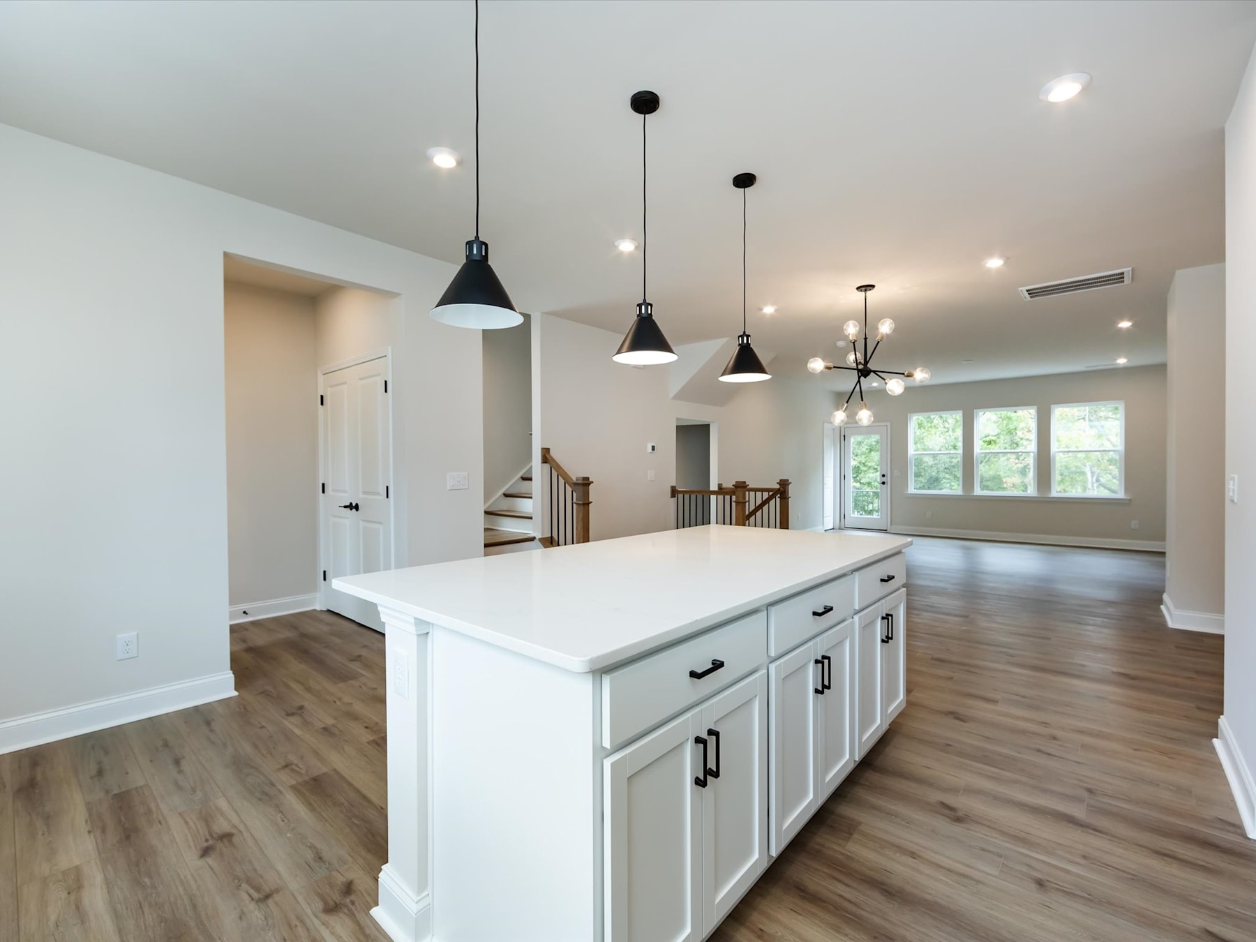 A modern, open-concept kitchen with a large white island, pendant lighting, and hardwood floors, leading into a living area with a staircase visible in the background.