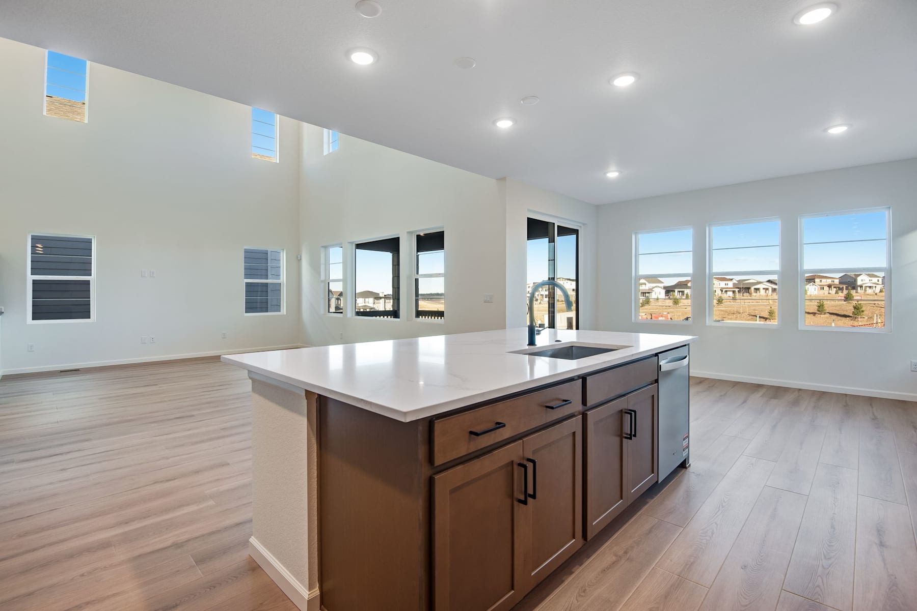 A modern, open-concept kitchen with a large island, wooden cabinets, and large windows overlooking a residential neighborhood in the background.