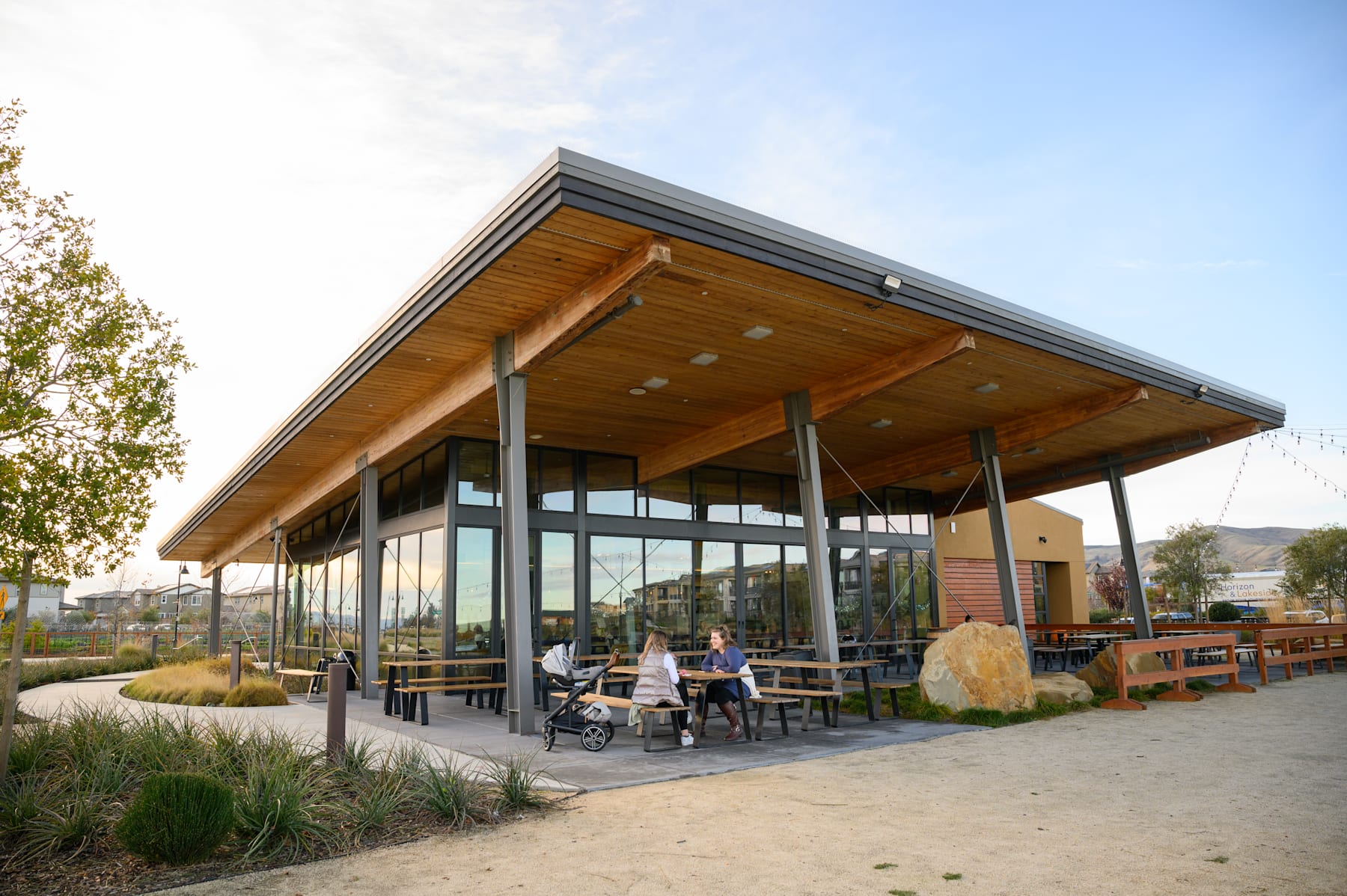 A modern, wooden-framed building with a large covered patio area, surrounded by landscaping and outdoor seating, set against a backdrop of a clear sky.