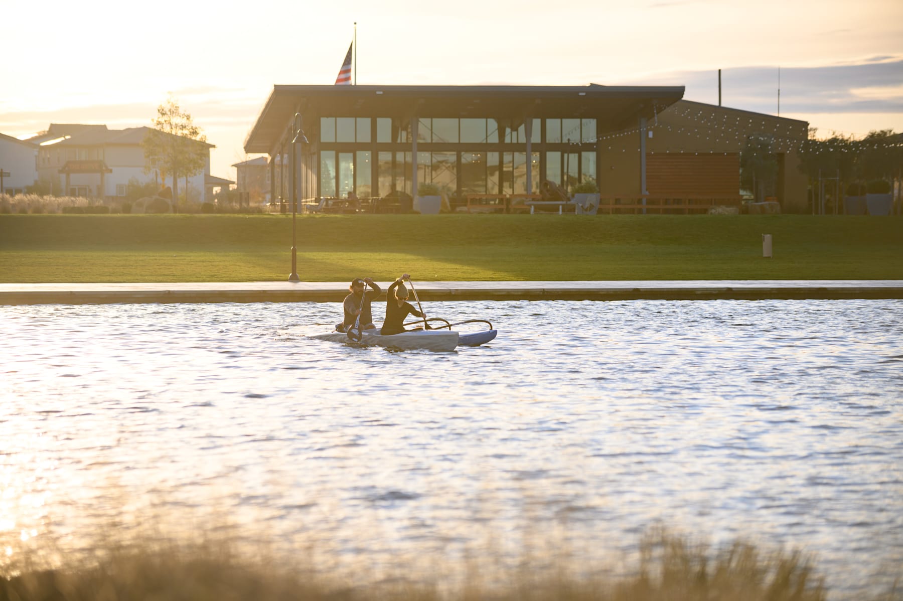 A small boat floats on a calm body of water, with a modern building and lush greenery visible in the background.