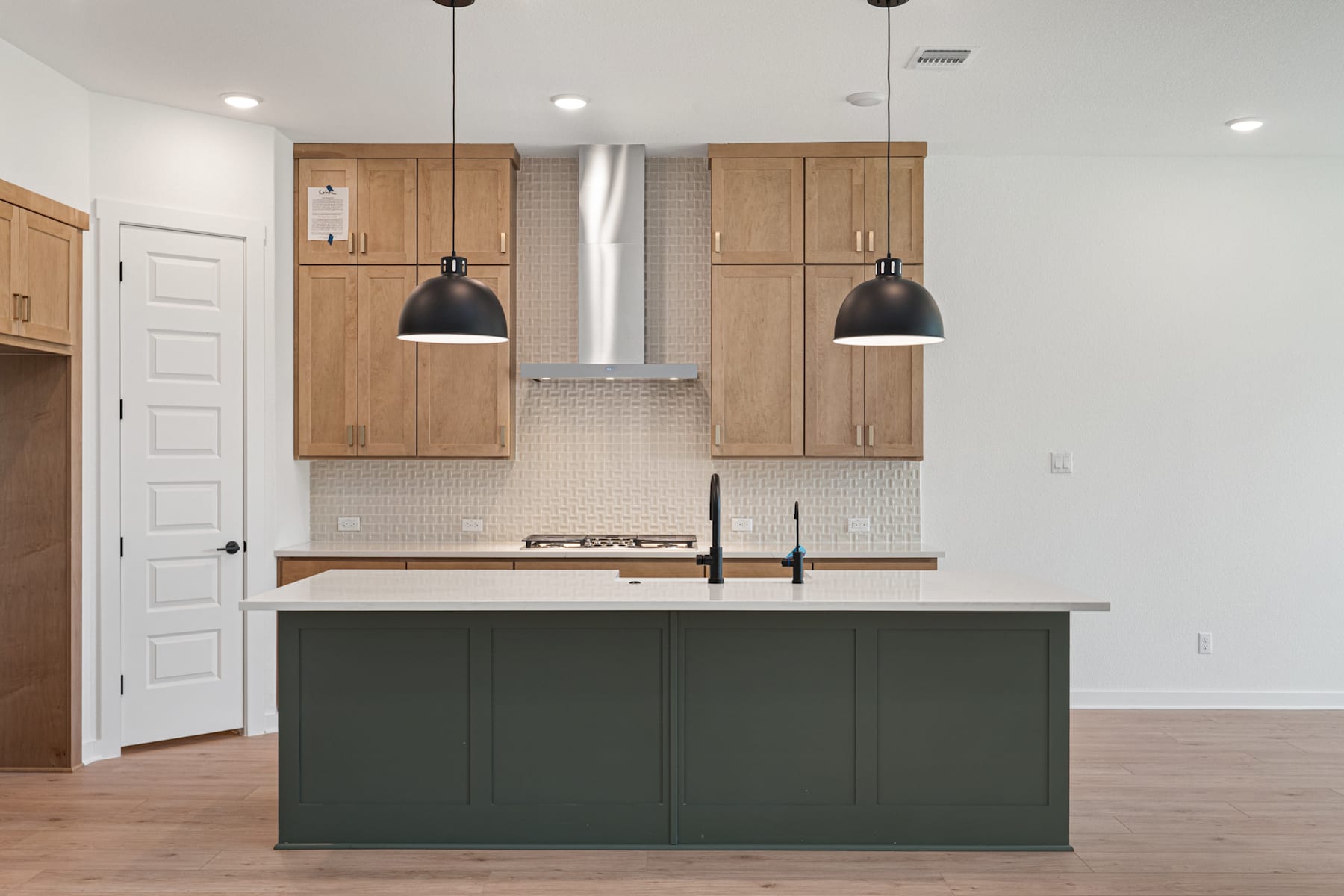 A modern kitchen with a green island, wooden cabinets, and pendant lights, set against a light-colored wall and hardwood flooring.