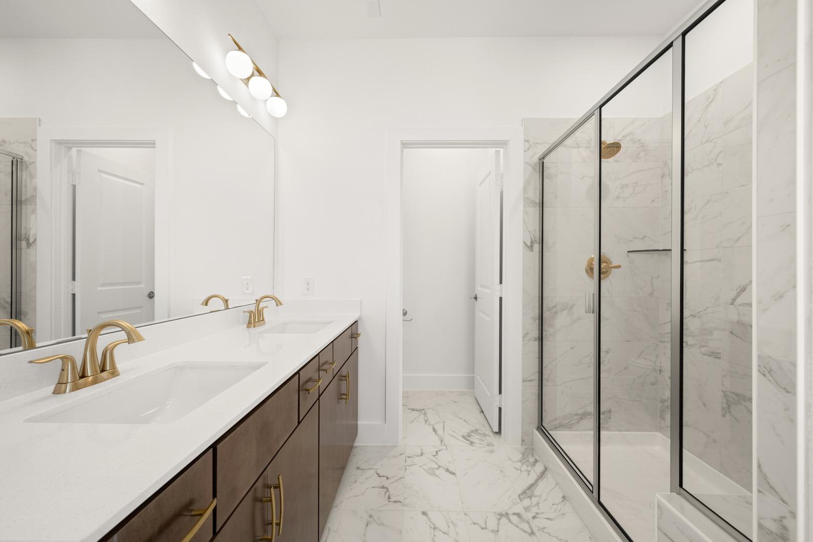 A modern and spacious bathroom with a marble-topped vanity, gold-toned fixtures, and a large glass-enclosed shower area.