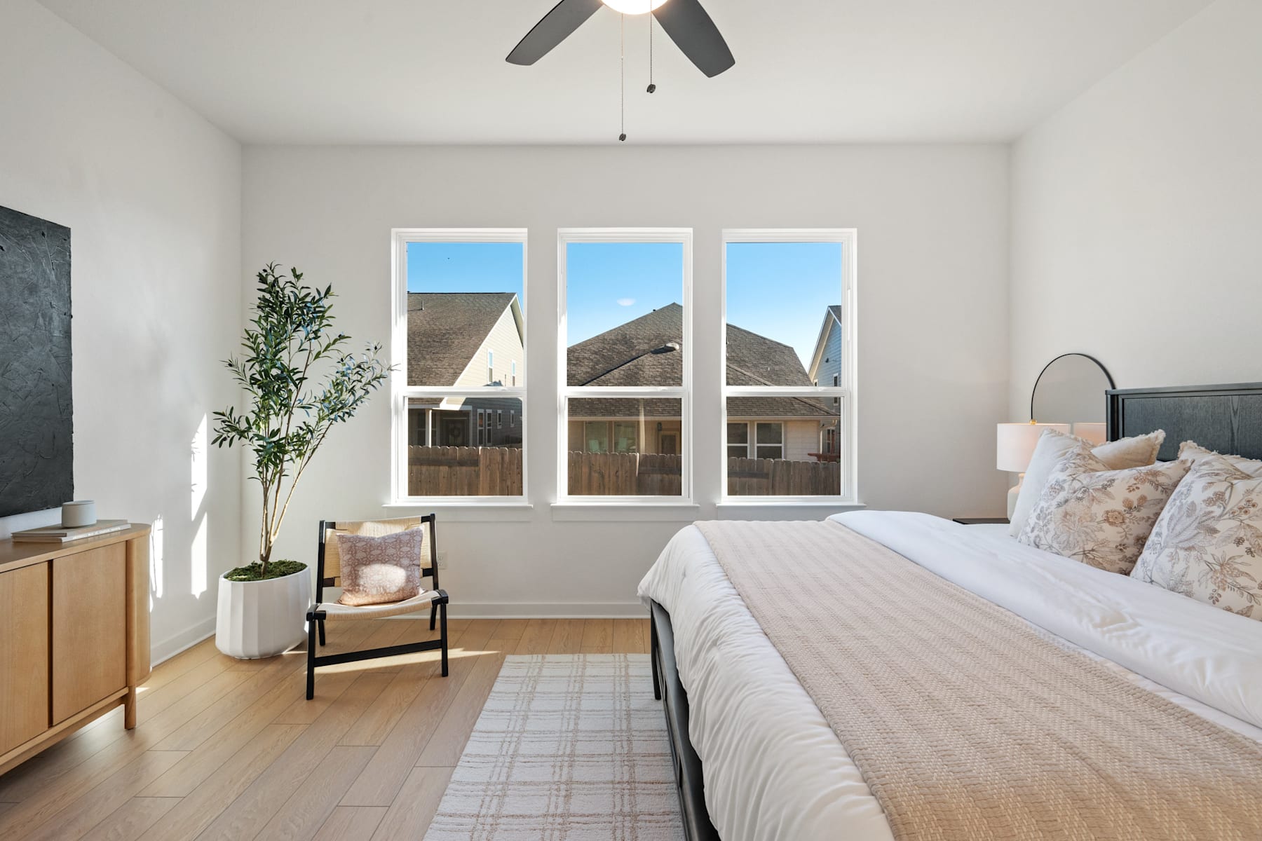 A cozy and well-lit bedroom with large windows overlooking a scenic outdoor landscape, featuring a comfortable bed, a potted plant, and a wooden dresser.
