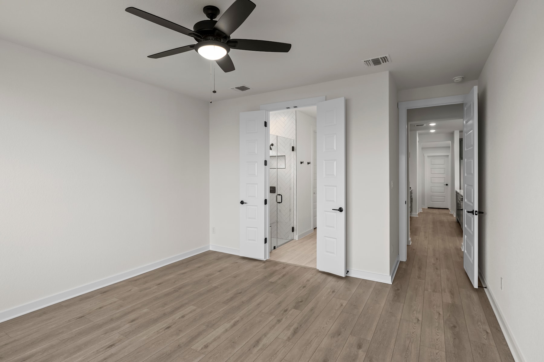 A spacious, minimalist room with a ceiling fan, white walls, and a wooden floor, leading to a hallway.