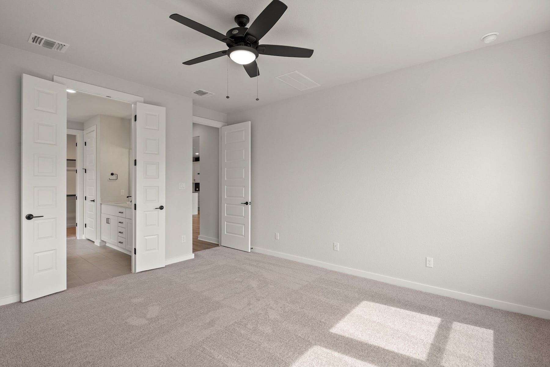 A spacious, minimalist bedroom with white doors, a ceiling fan, and a carpeted floor bathed in natural light.