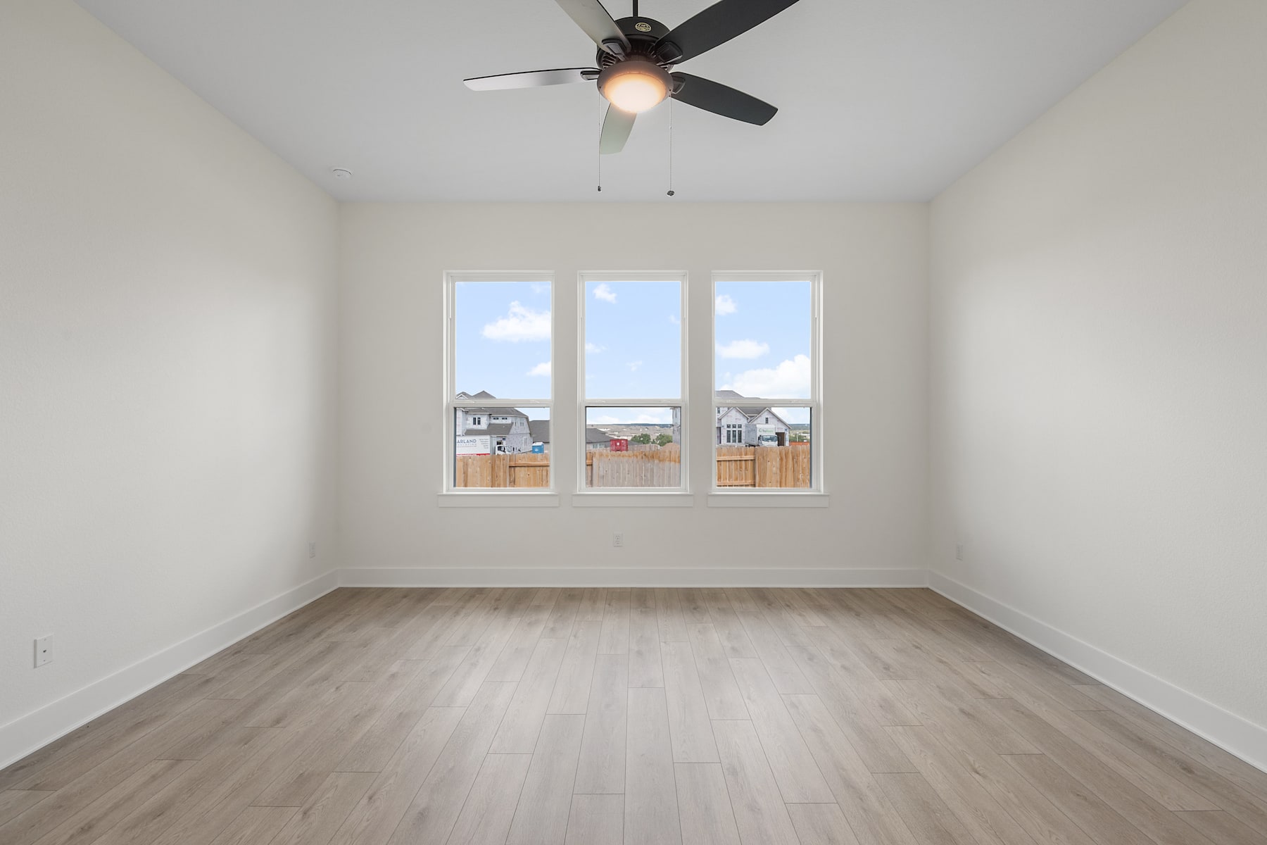 An empty, bright room with a ceiling fan and large windows overlooking a residential neighborhood.