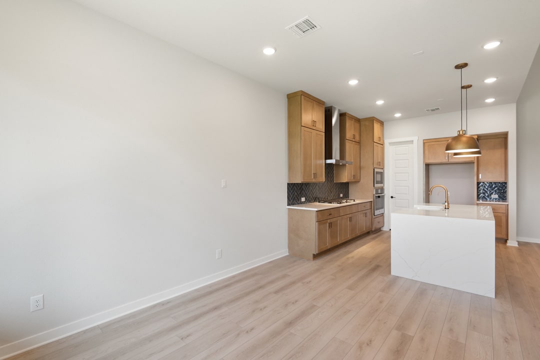 A modern, open-concept kitchen with light-colored wood cabinets, a white countertop, and hardwood flooring extends into a bright, airy living space.