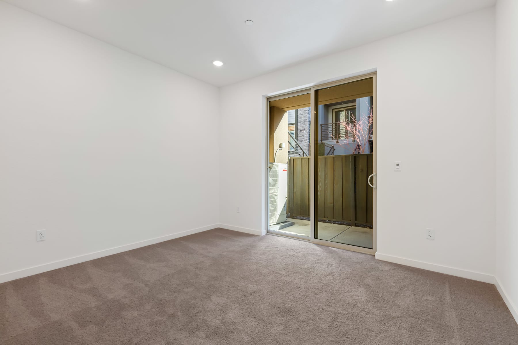A spacious, minimalist room with a sliding glass door leading to an outdoor area, featuring a neutral-toned carpet and white walls.