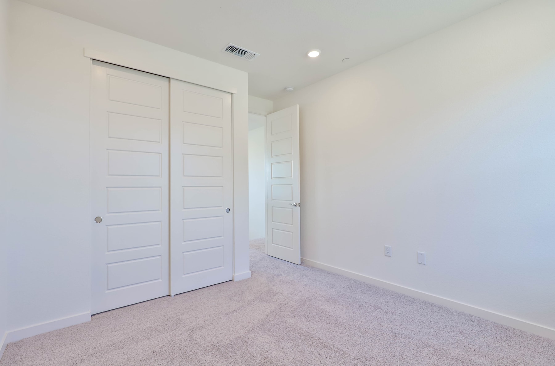 A simple, minimalist bedroom with white walls, a closed door, and a carpeted floor.
