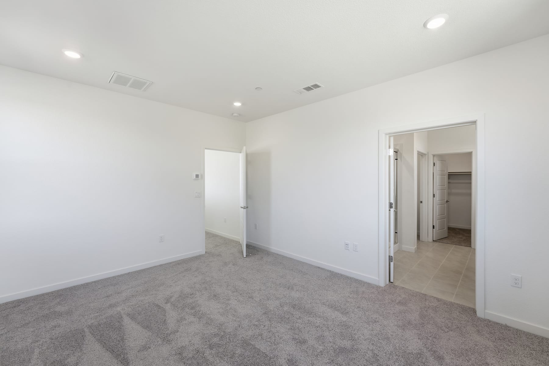 A spacious, empty room with white walls, gray carpeting, and recessed lighting fixtures on the ceiling.