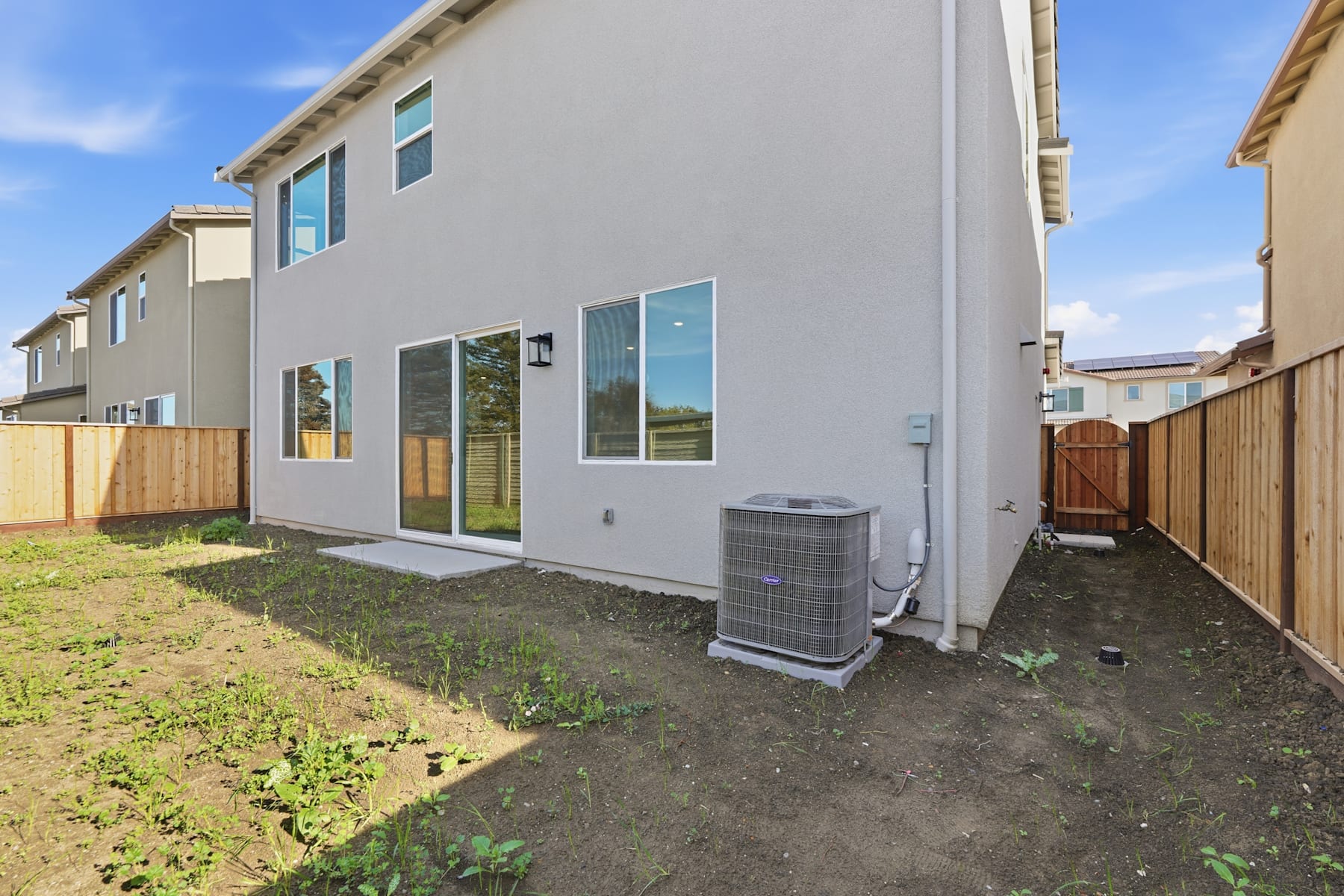 A modern, two-story residential building with a grassy yard and a central air conditioning unit in the foreground, set against a clear blue sky.
