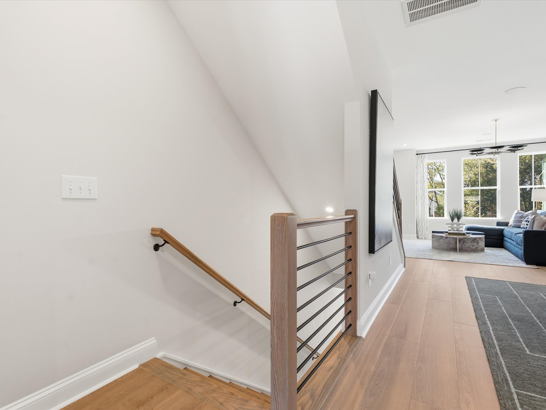 A modern, open-concept interior with a wooden staircase railing leading to a living room area with a large window overlooking the outdoors.