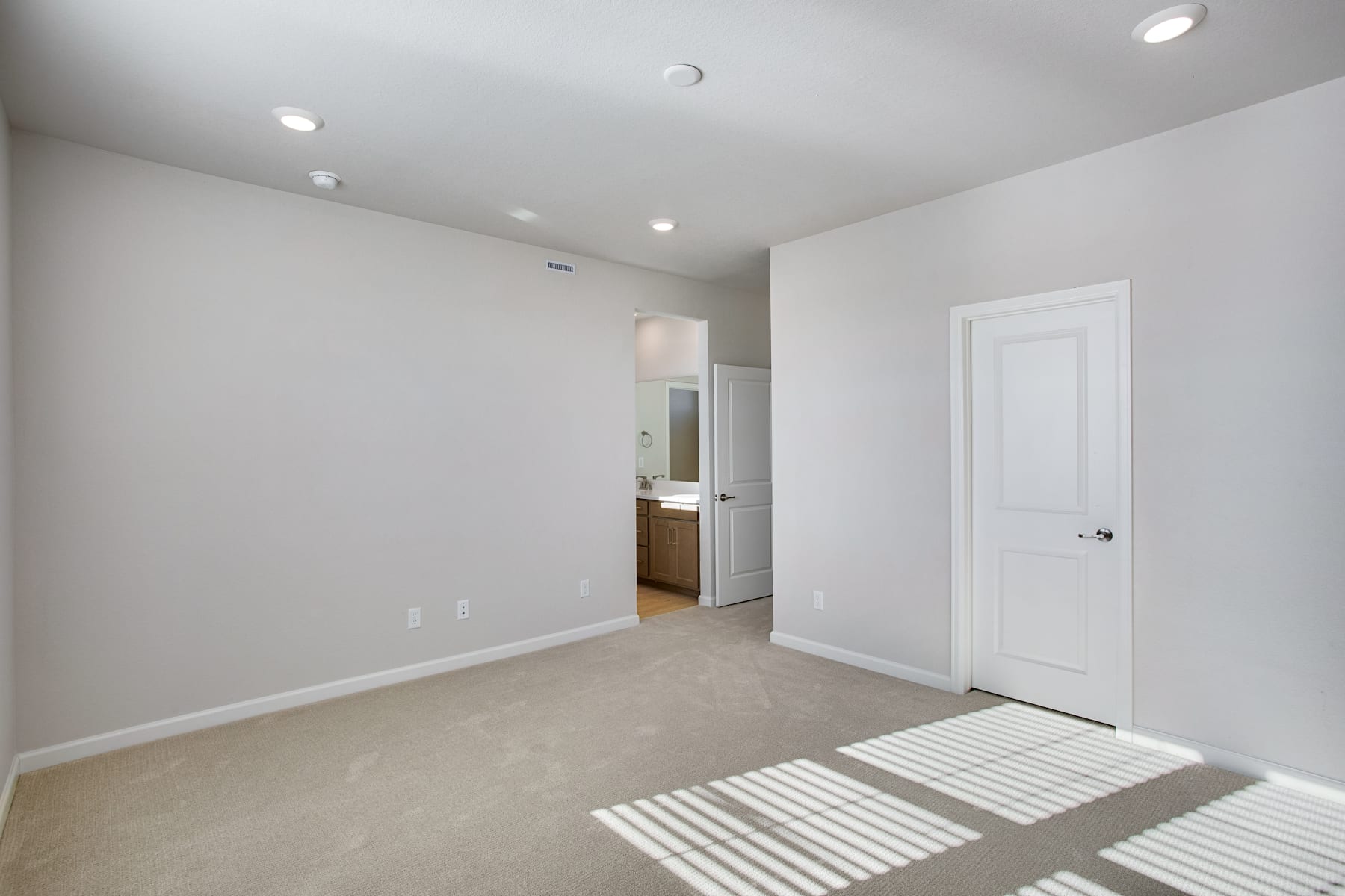 A spacious, well-lit room with white walls, a carpeted floor, and a partially visible doorway leading to another room.