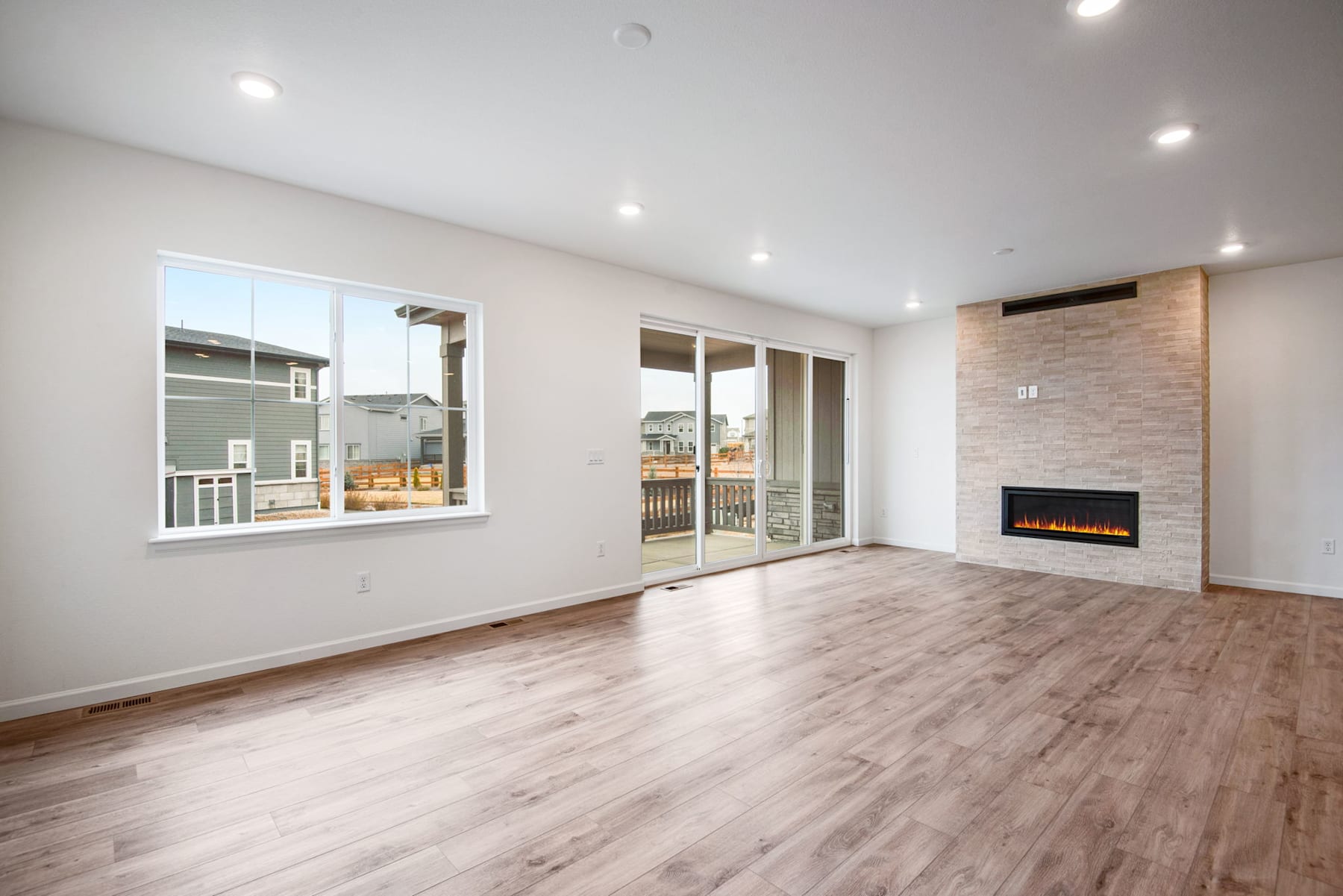 A spacious and modern living room with a fireplace, hardwood floors, and large windows overlooking a residential neighborhood.