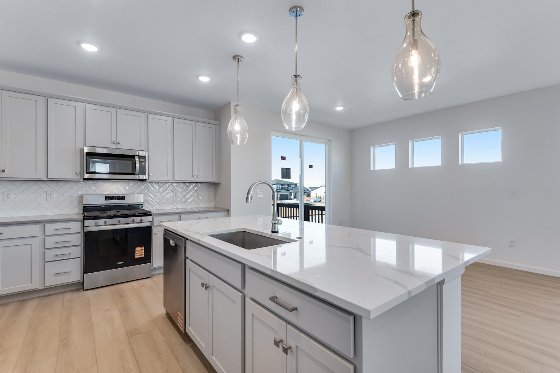 A modern, well-lit kitchen with white cabinets, stainless steel appliances, and a large island with a white countertop, set against a backdrop of large windows and natural light.