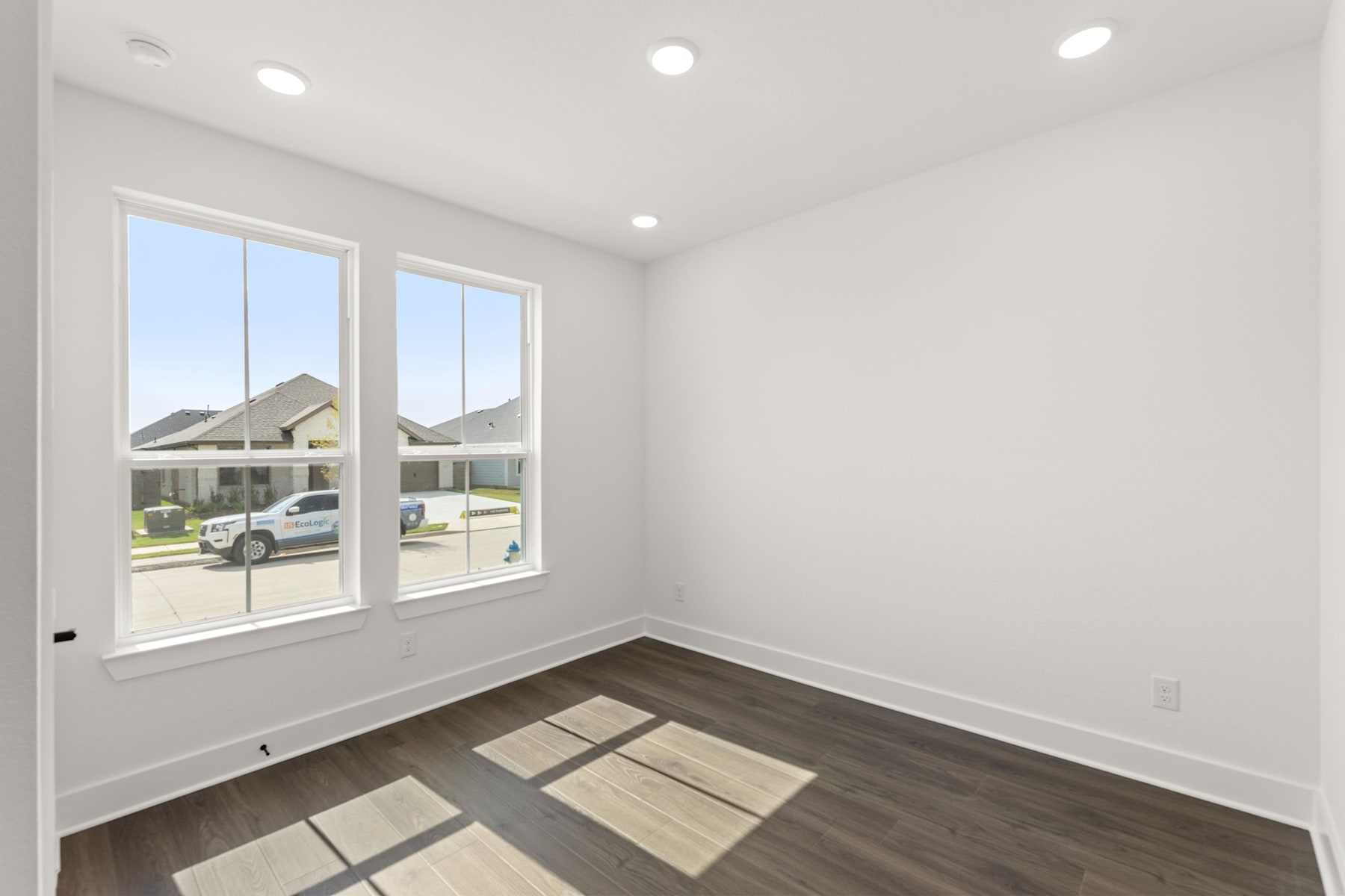 A bright, empty room with large windows overlooking a residential neighborhood, featuring a hardwood floor and recessed lighting.