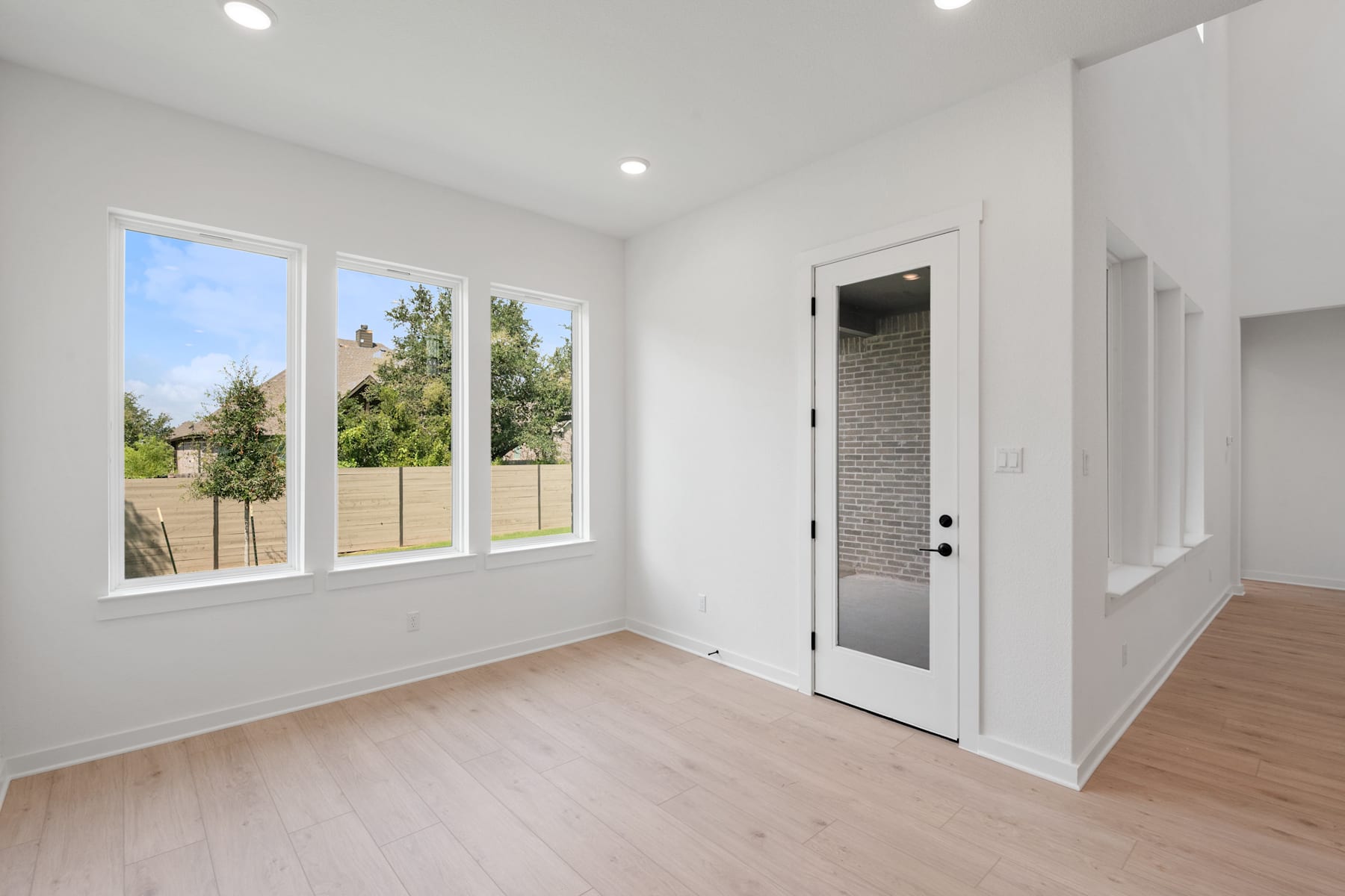 A bright and airy room with large windows overlooking a lush outdoor landscape, featuring a wooden floor and a white door leading to another area.