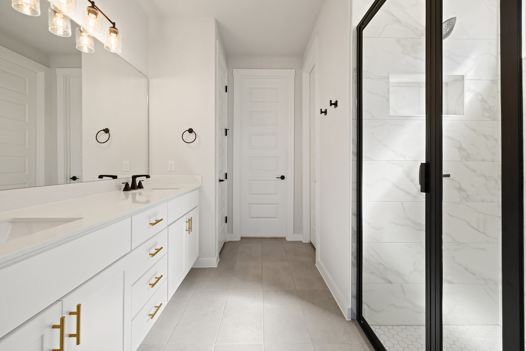A bright and spacious bathroom with a long vanity, white cabinets, and gold hardware, leading to a hallway with white walls and doors.