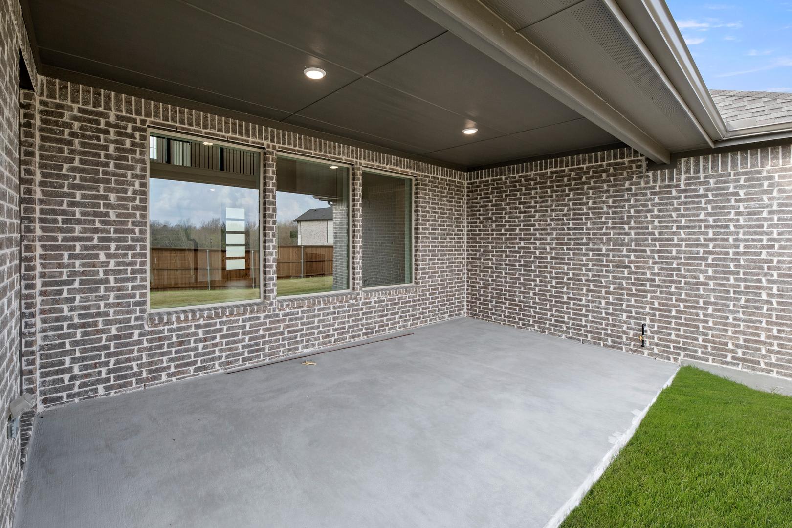 A covered porch with a brick exterior, concrete floor, and windows overlooking a grassy yard in the background.