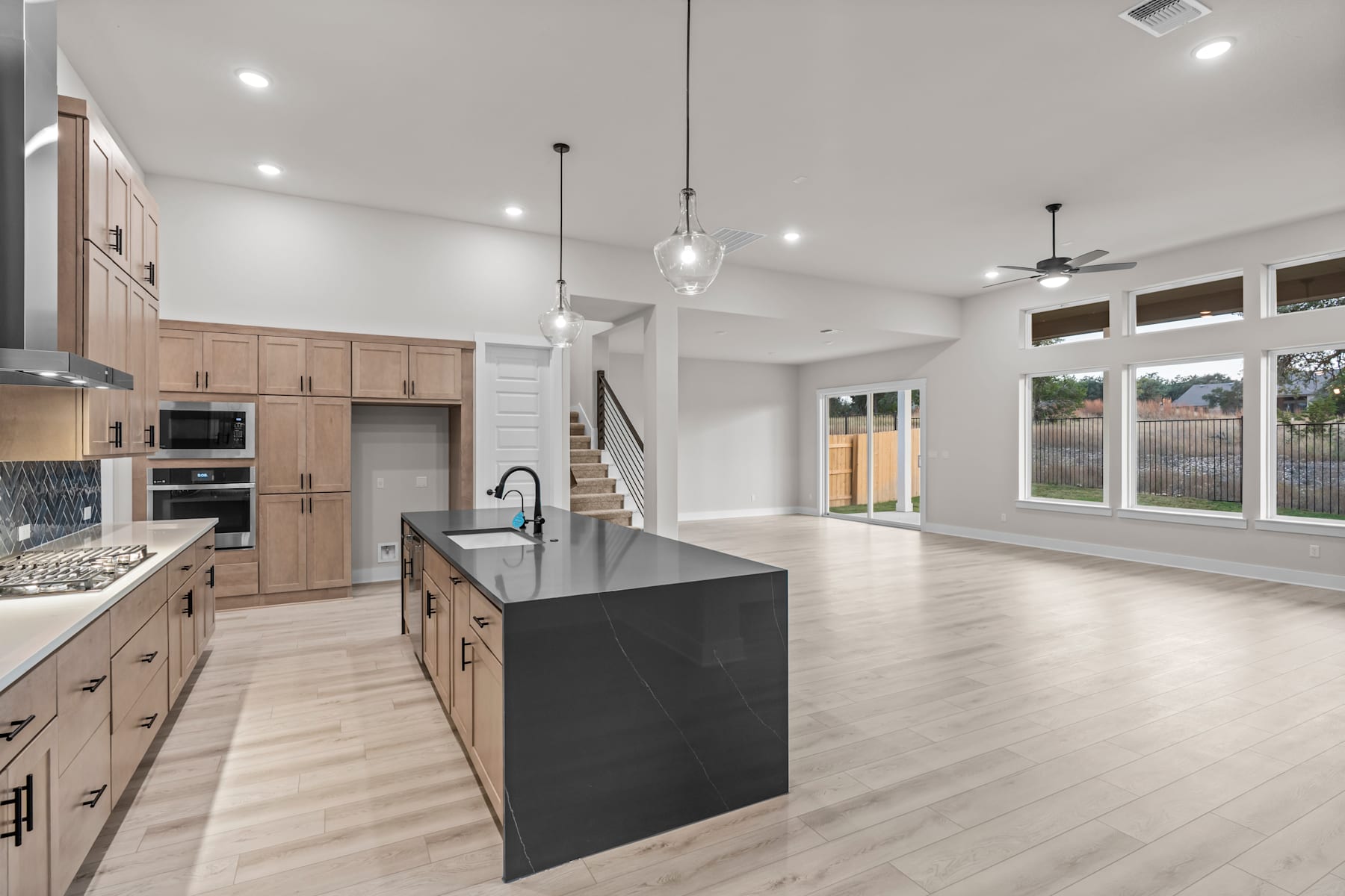 A modern, open-concept kitchen with light wood cabinets, a large black island, and hardwood floors, surrounded by large windows that let in natural light.
