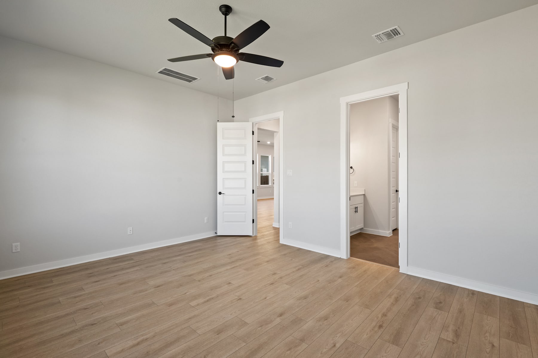 An empty room with a ceiling fan, wooden floors, and white walls, with a doorway leading to another room.