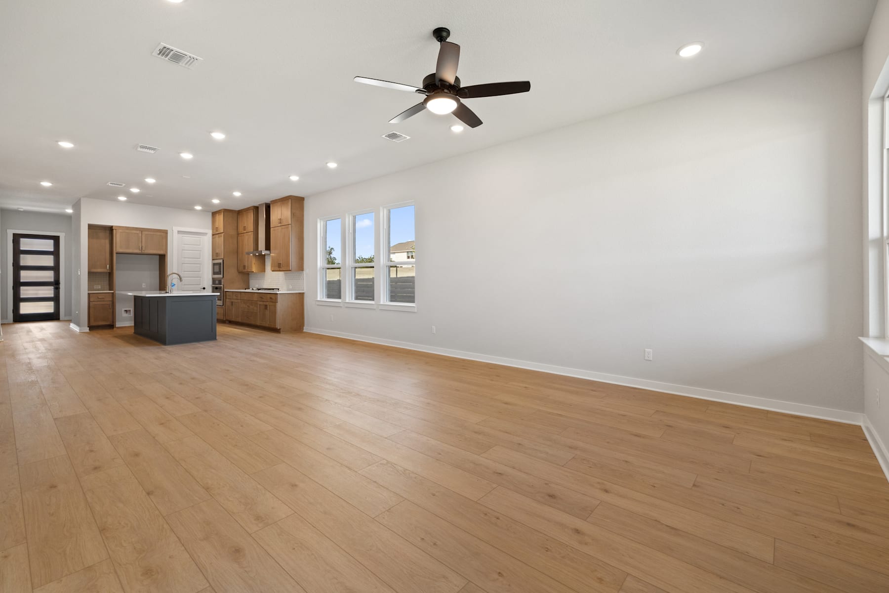 A spacious and well-lit room with a ceiling fan, wooden floors, and a kitchen visible in the background.