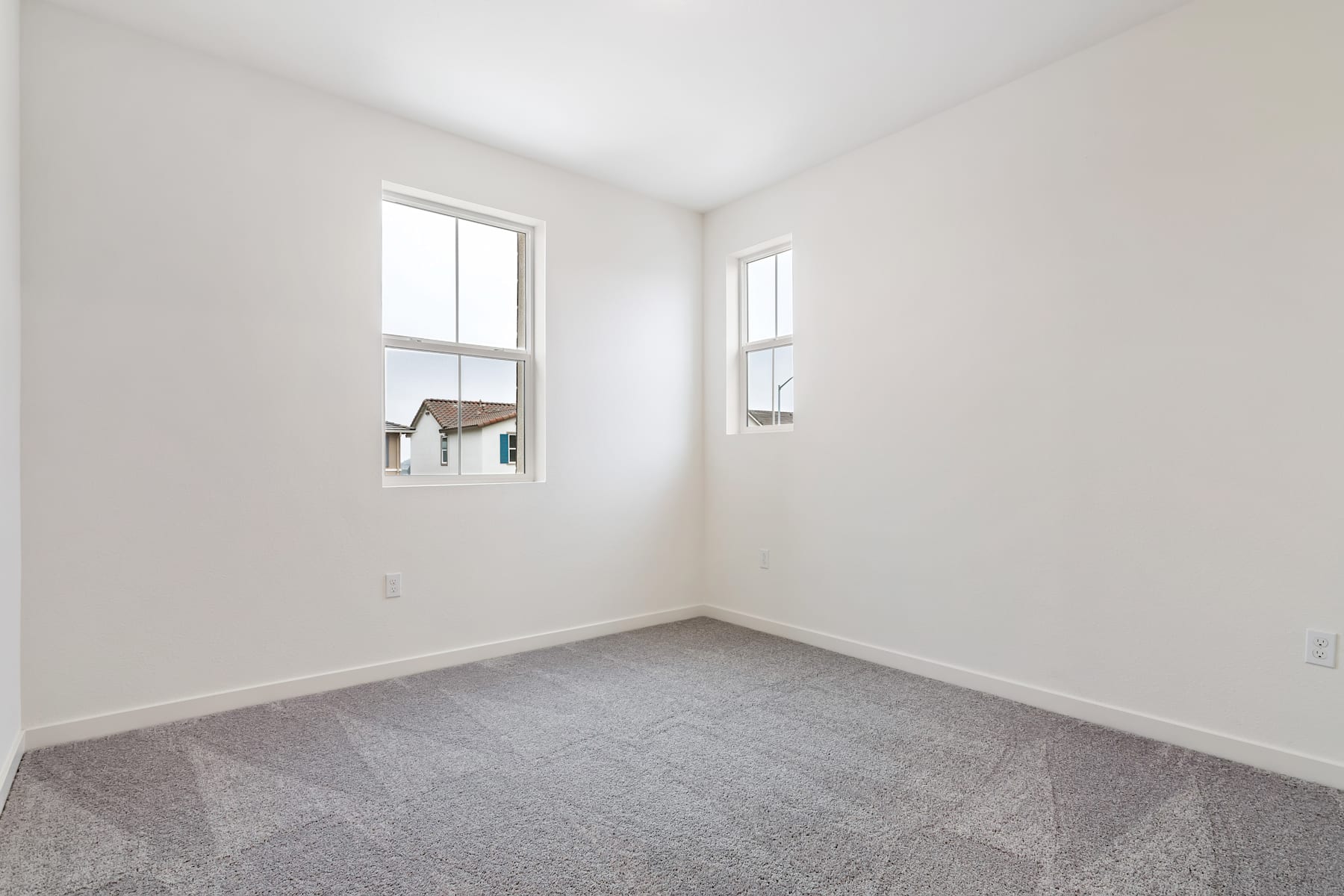 A simple, minimalist bedroom with white walls, a gray carpeted floor, and two windows providing natural light.
