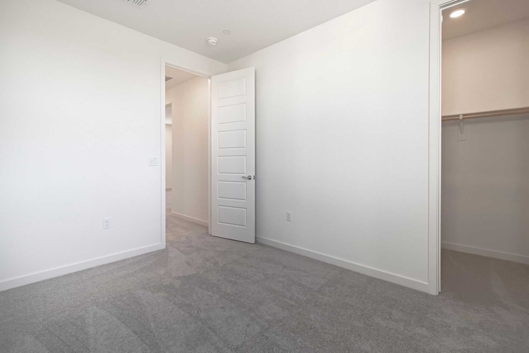 A spacious, minimalist room with white walls, a closed door, and a gray carpeted floor.