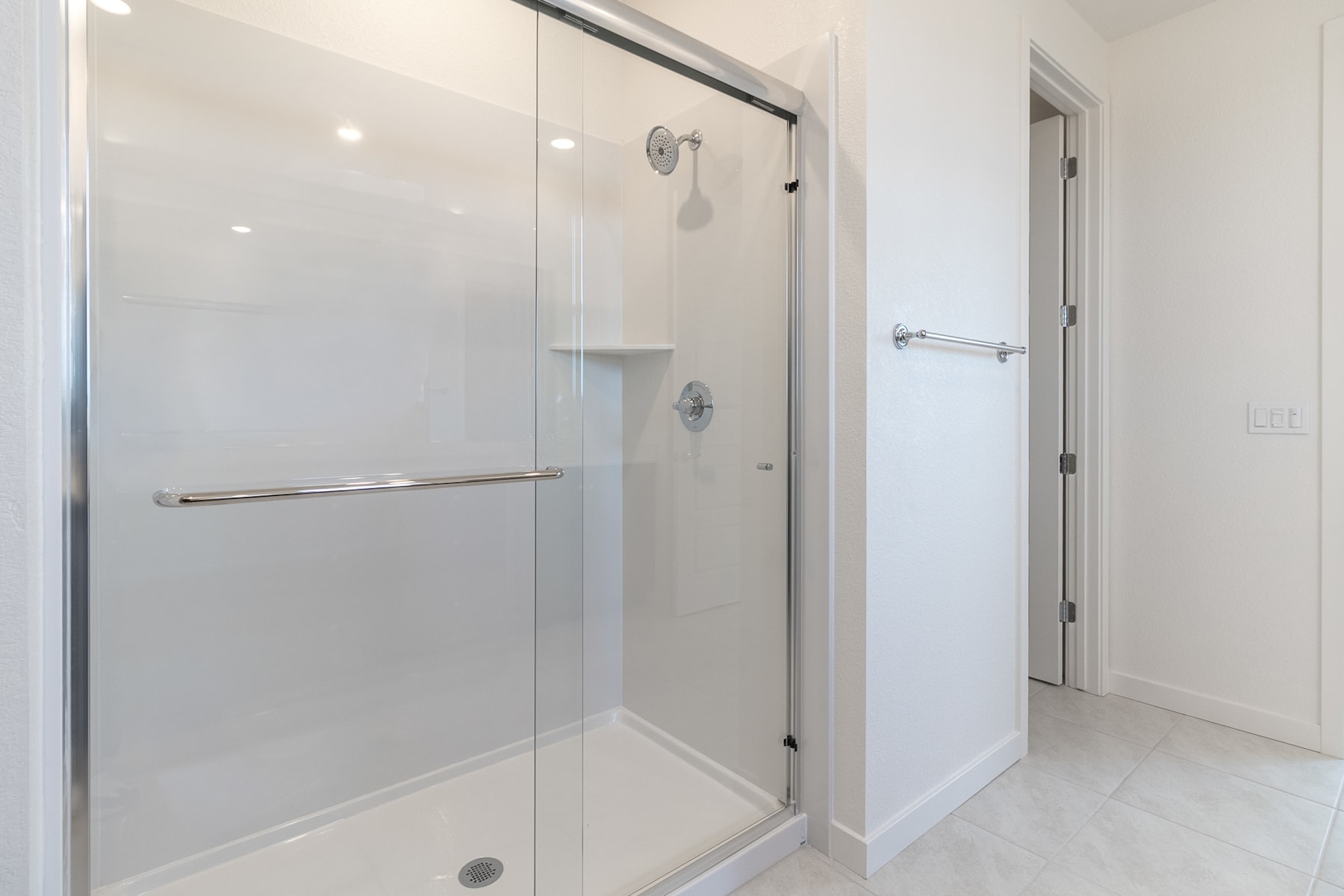 A modern, minimalist bathroom with a glass shower enclosure, a white tiled floor, and a closed door in the background.
