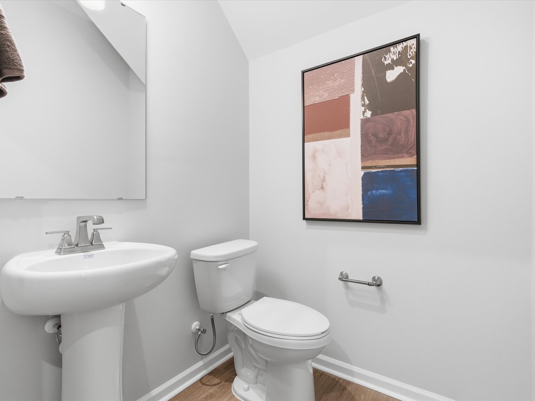 A modern, minimalist bathroom with a white pedestal sink, toilet, and a framed abstract artwork on the wall.