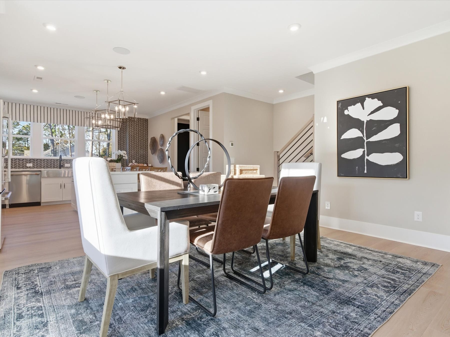 A modern and stylish dining room with a large wooden table, white chairs, and a chandelier overhead, set against a backdrop of a kitchen area with stainless steel appliances.