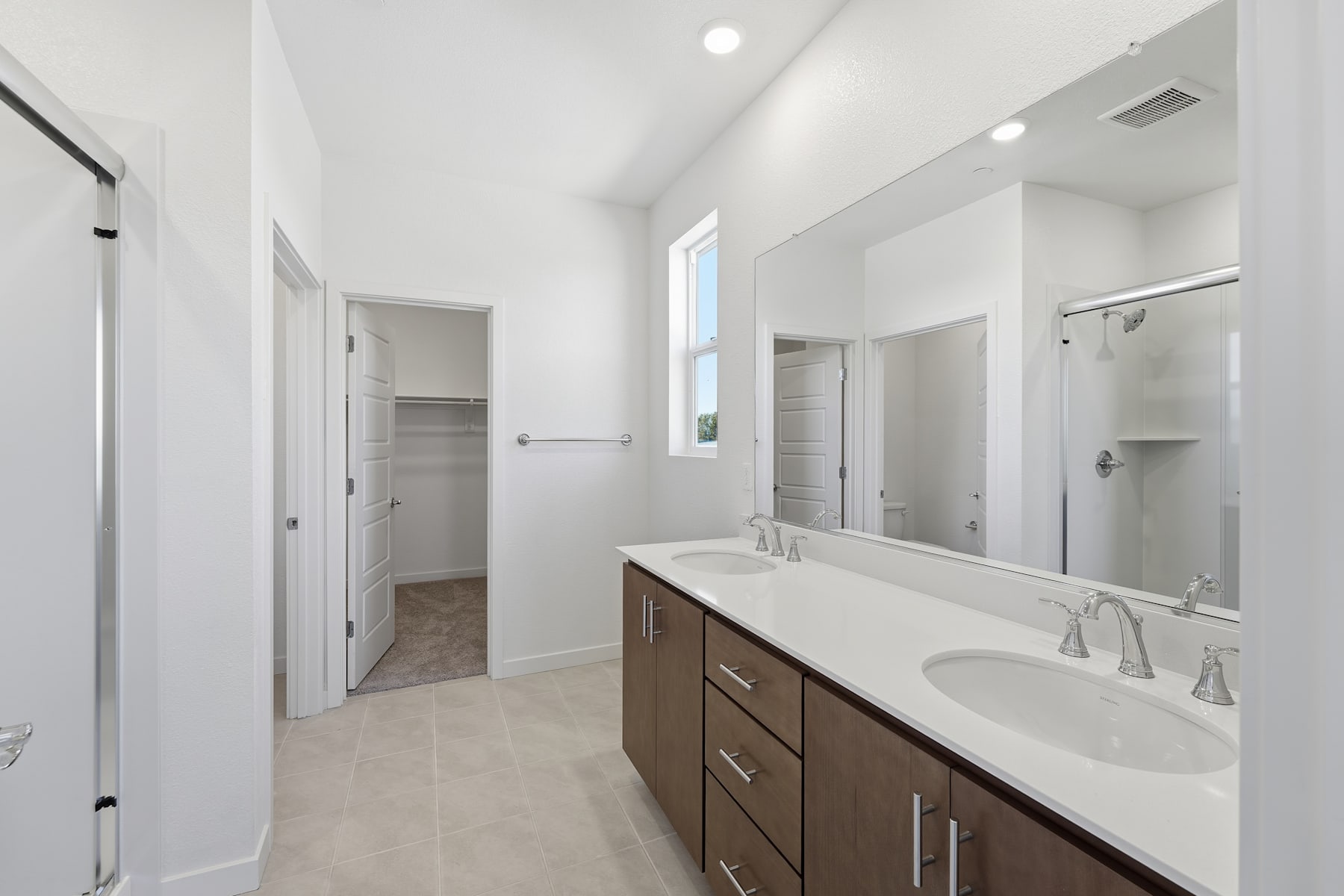 A spacious and modern bathroom with a large vanity, double sinks, and a mirror taking up the majority of the wall space. The room has a clean, minimalist aesthetic with white walls and tile flooring.