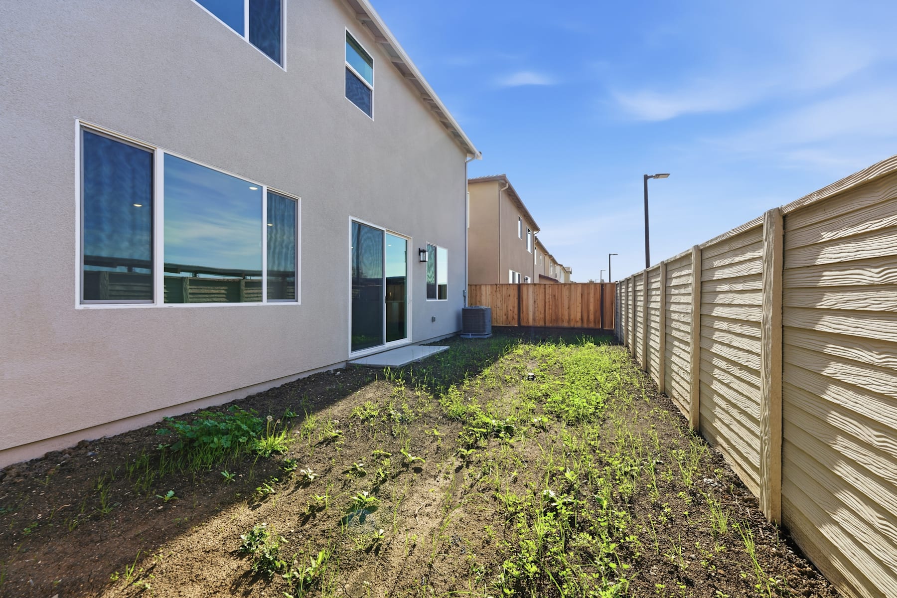 The image shows a narrow walkway between a residential building and a wooden fence, with a grassy area in the foreground and a clear blue sky in the background.