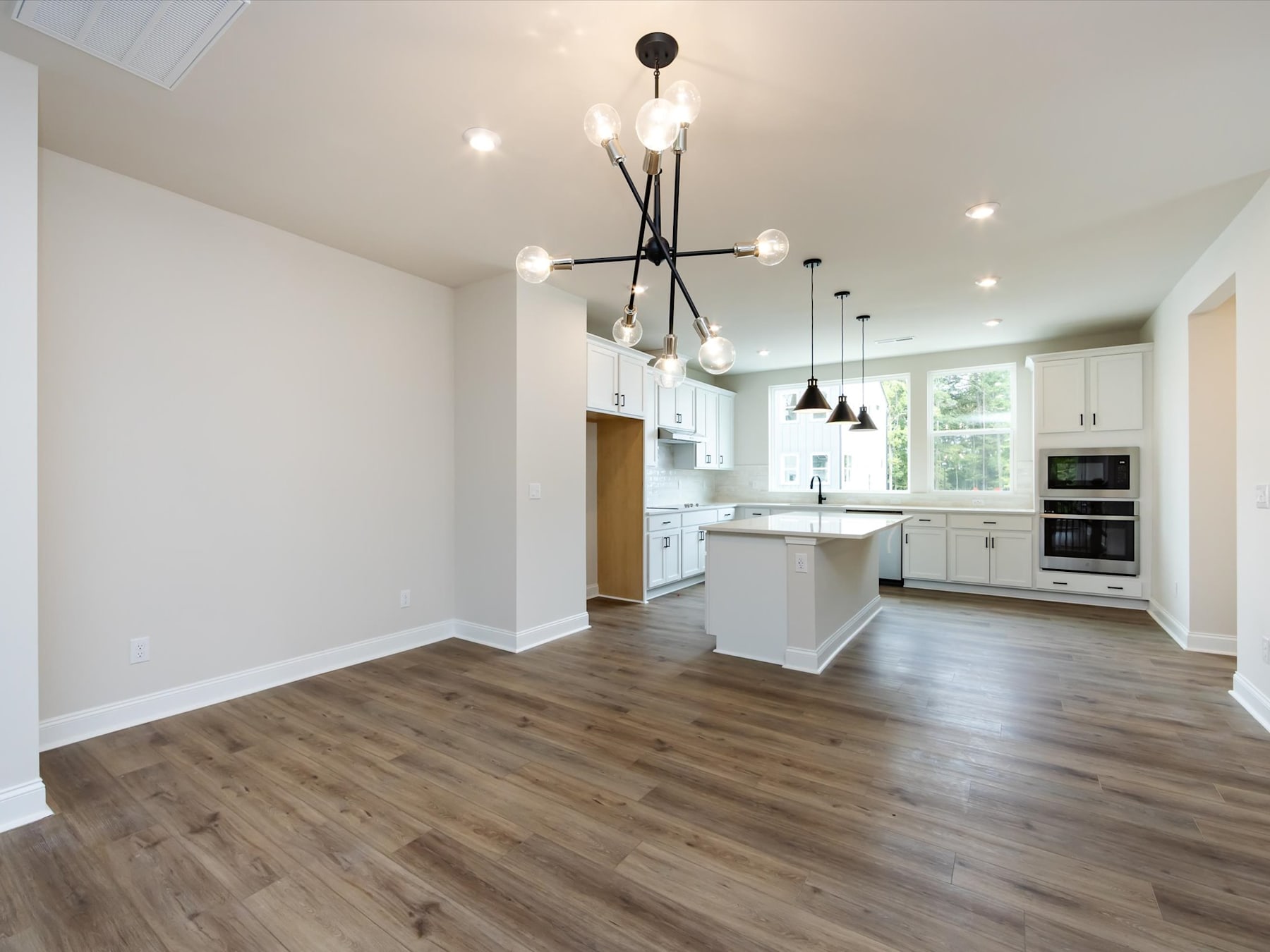 A modern, open-concept kitchen and living room with white walls, hardwood floors, and a stylish black chandelier hanging overhead.