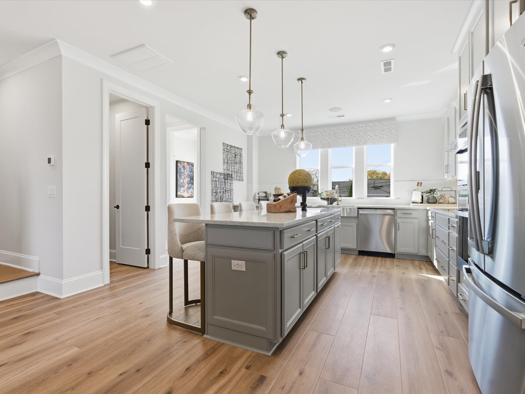 A modern, well-lit kitchen with a gray island, stainless steel appliances, and pendant lighting fixtures, set against a backdrop of white walls and hardwood floors.
