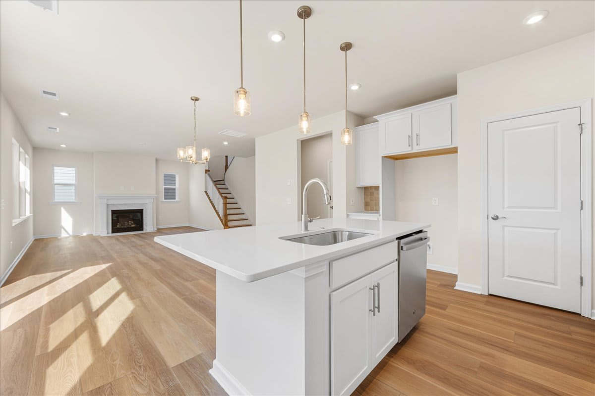 A modern, open-concept kitchen with white cabinets, a large island, and pendant lighting fixtures, set against a backdrop of hardwood floors and a staircase.