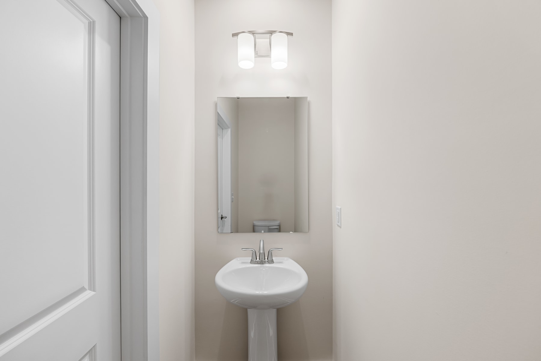 A small, minimalist bathroom with a white pedestal sink and a light fixture above it, set against a plain white wall.