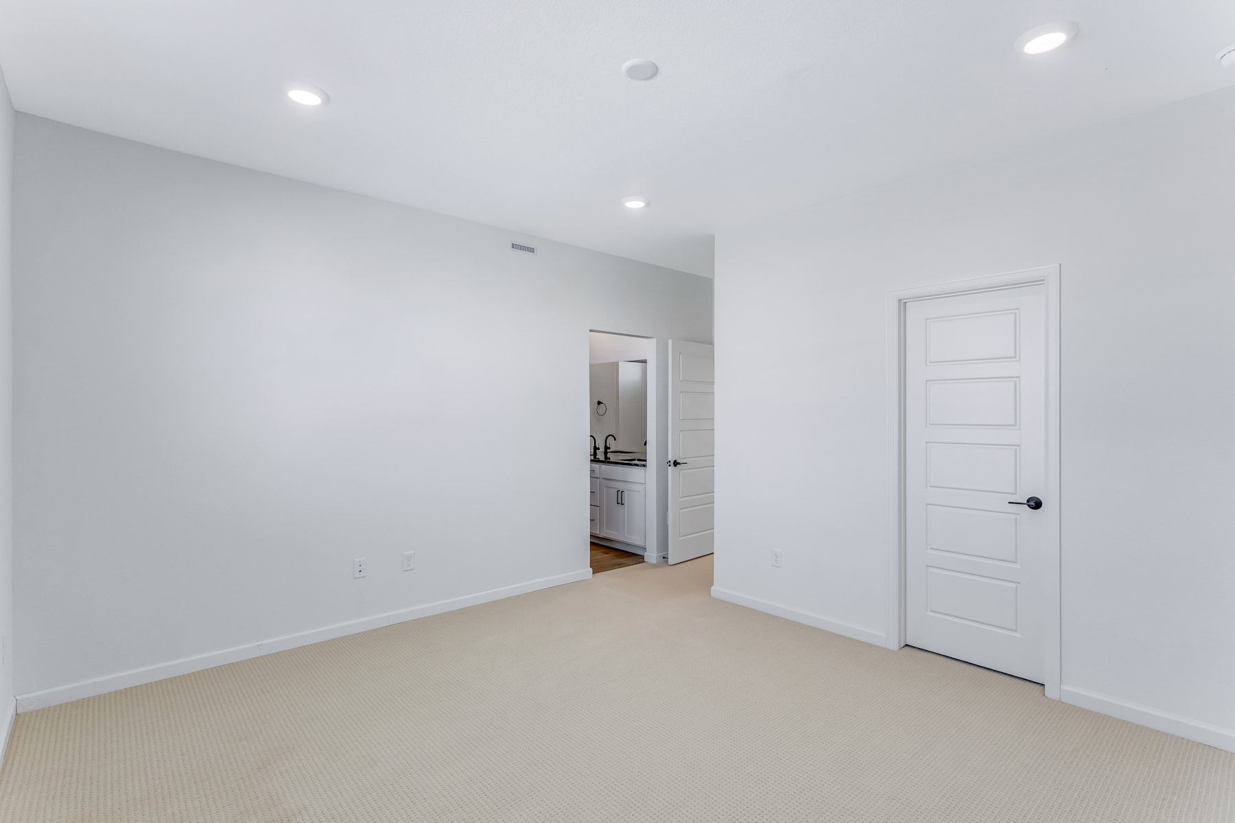 A spacious, minimalist room with white walls, a white door, and recessed lighting in the ceiling.