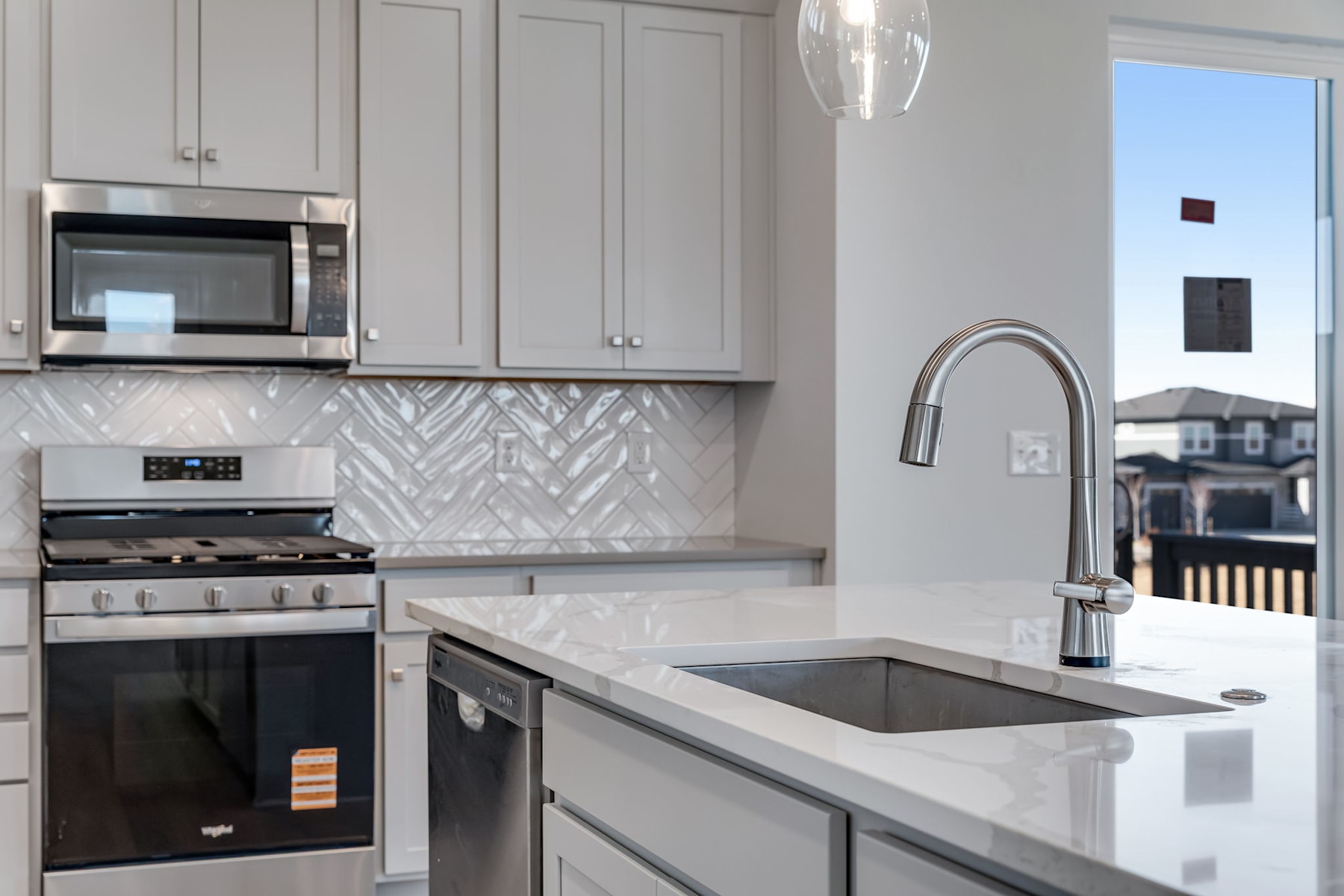A modern, well-equipped kitchen with gray cabinets, a stainless steel oven and microwave, and a sleek, white countertop with a sink and faucet. The background includes a window overlooking what appears to be a residential neighborhood.