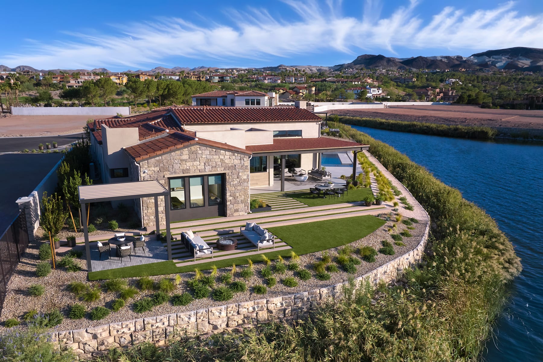 A modern, stone-clad house with a tiled roof sits on a hillside, surrounded by lush greenery and overlooking a body of water, with a mountainous landscape in the background.