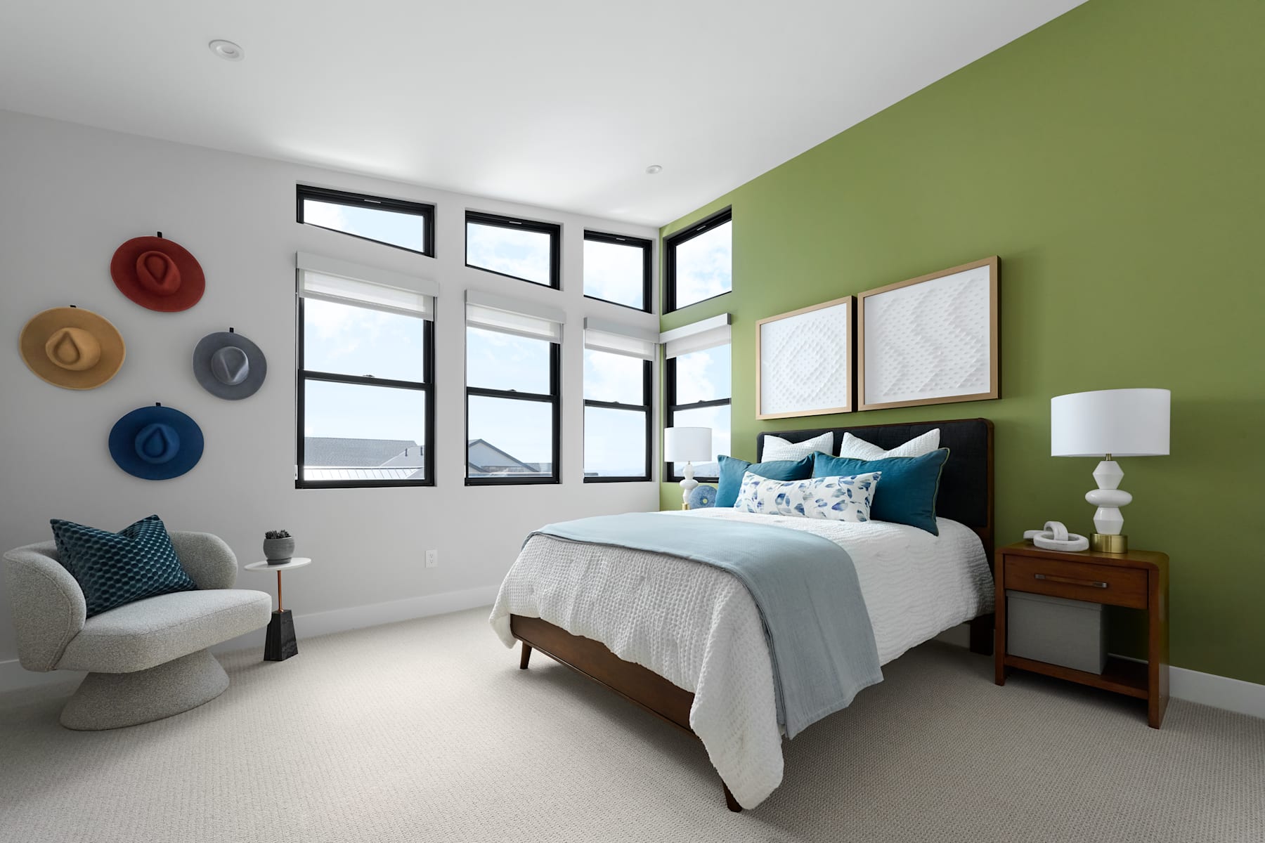 A cozy and modern bedroom with a green accent wall, large windows, and various decorative elements such as hats and framed artwork adorning the walls.