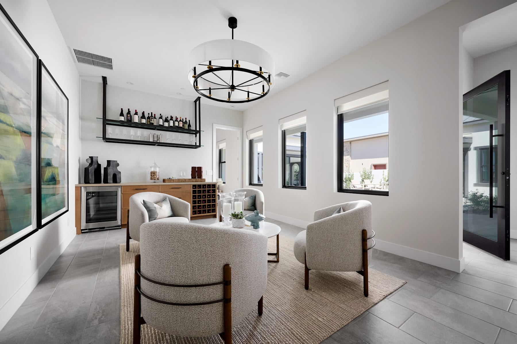 A modern and stylish dining room with a chandelier, comfortable chairs, and a wine rack on the wall, surrounded by large windows and a neutral color scheme.