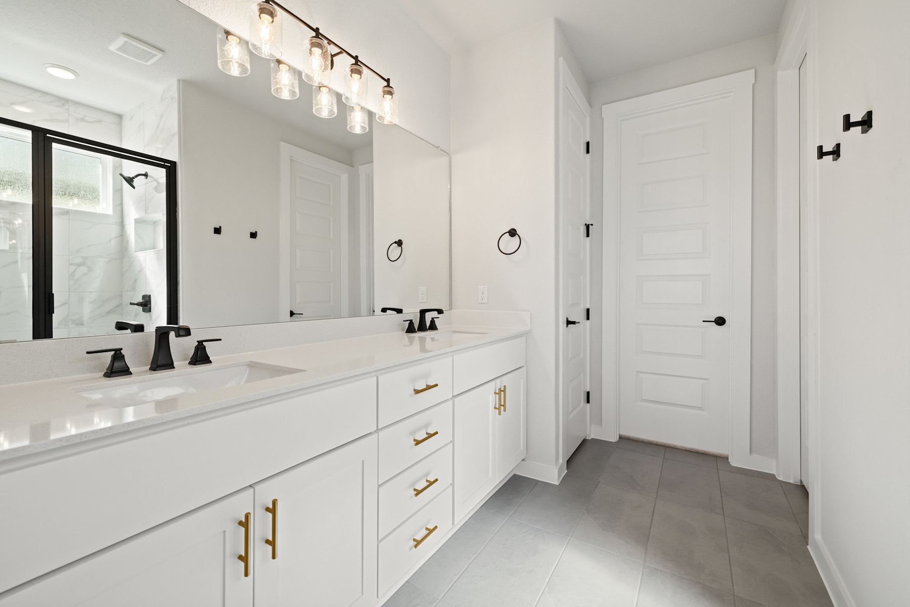 A modern, minimalist bathroom with white cabinets, a double vanity, and a tiled floor. The room is well-lit with recessed lighting and wall sconces.