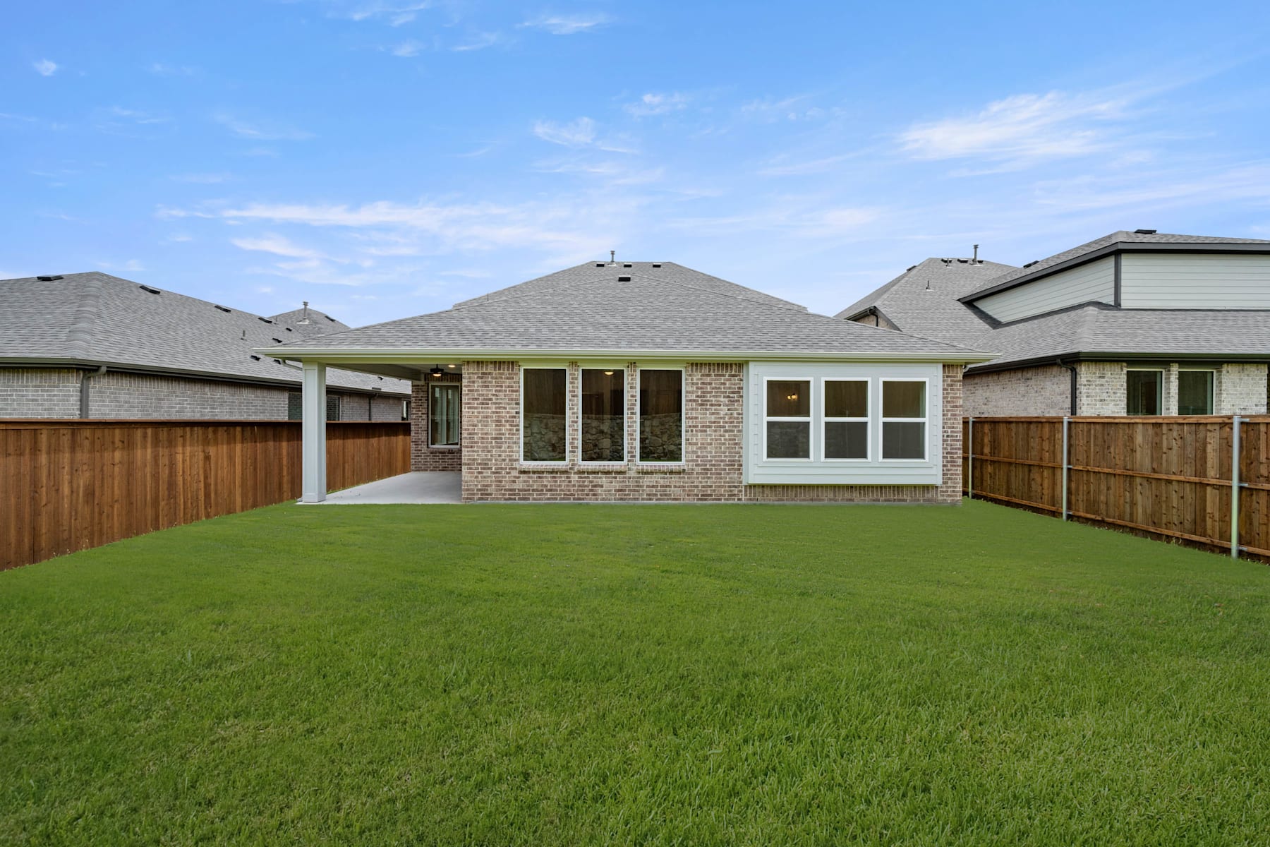 A well-manicured lawn in the foreground leads to a modern, single-story house with a brick exterior and a peaked roof in the background, set against a clear blue sky.