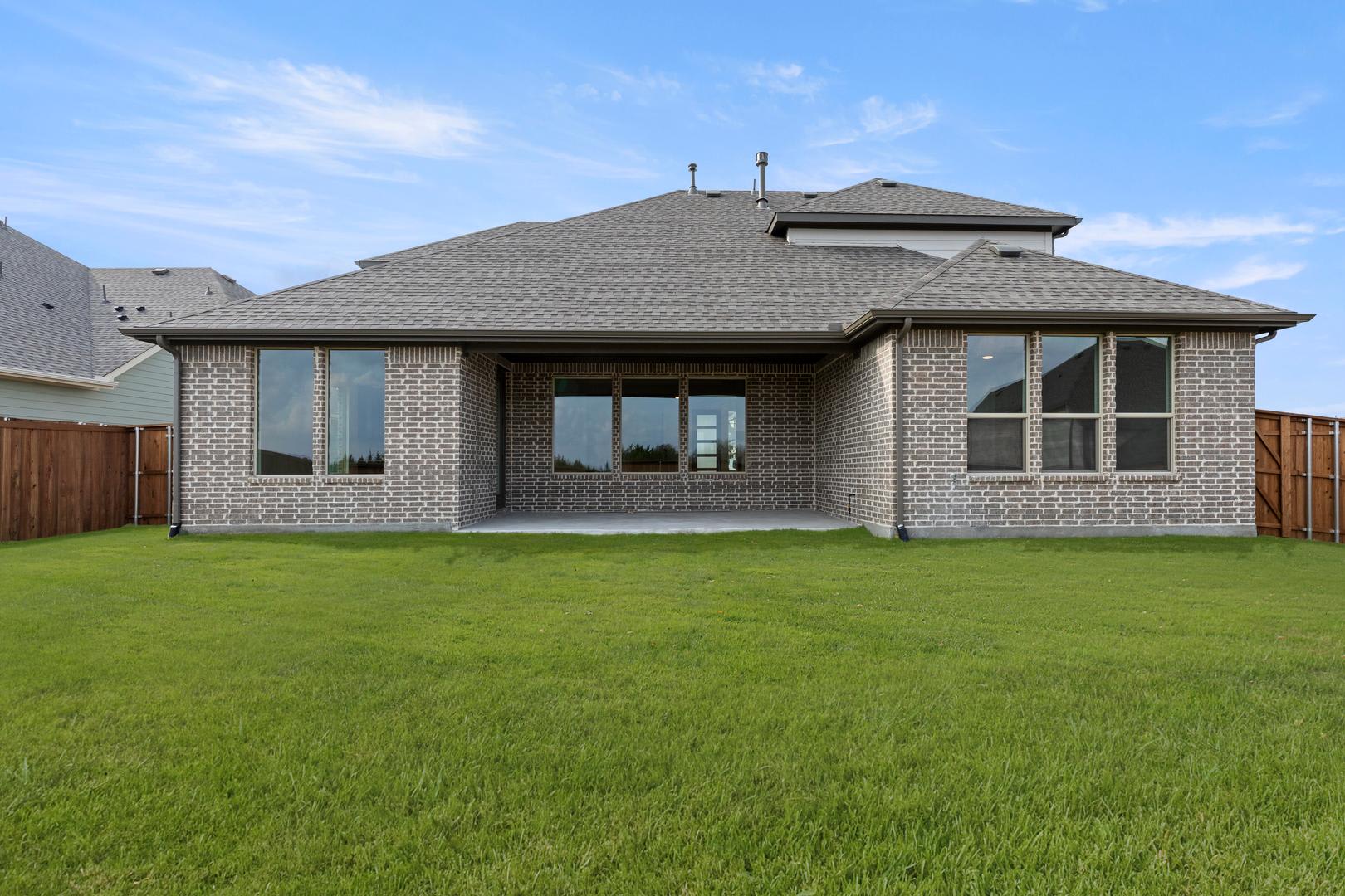A single-story brick house with a gabled roof sits on a lush green lawn, surrounded by a clear blue sky with scattered clouds.