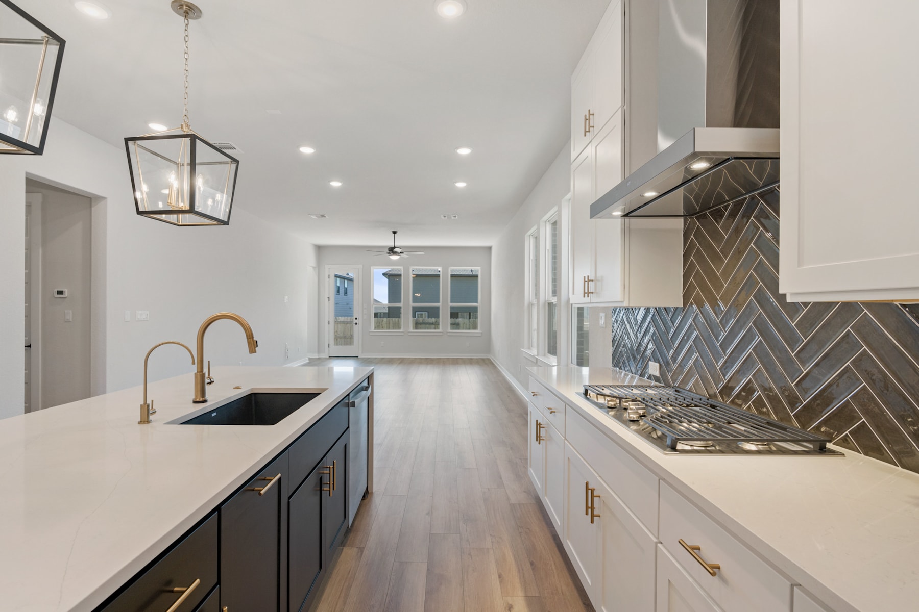 A modern, open-concept kitchen with sleek black cabinets, white countertops, and a herringbone-patterned backsplash, leading into a bright and airy hallway with hardwood floors.