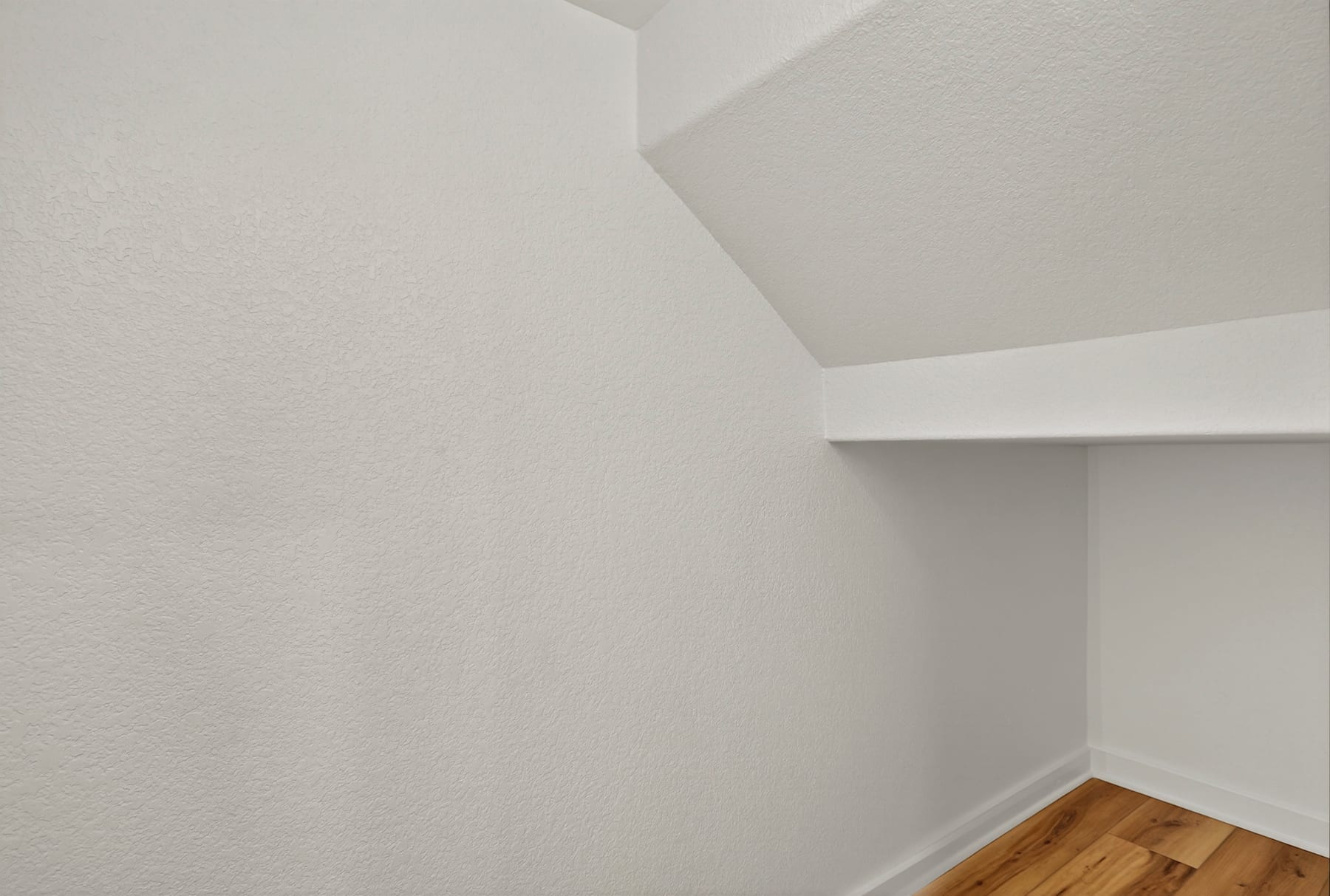 A simple, minimalist room with white walls and a wooden floor, featuring a recessed ceiling and a shelf or ledge in the corner.