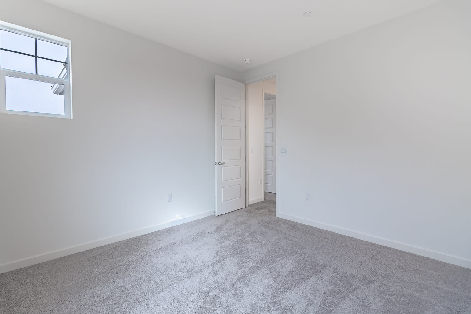 A simple, minimalist bedroom with white walls, a window, and a gray carpeted floor.