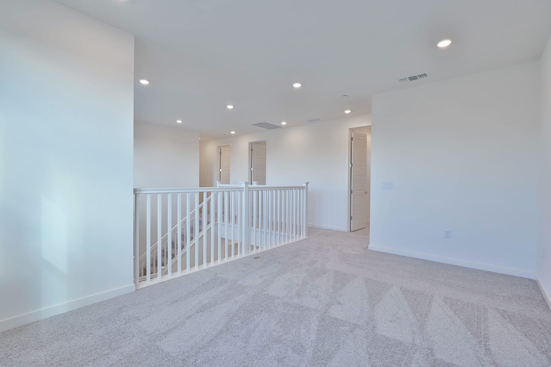 A spacious, well-lit hallway with a white railing leading to a doorway, featuring a neutral-toned carpet and recessed lighting in the ceiling.