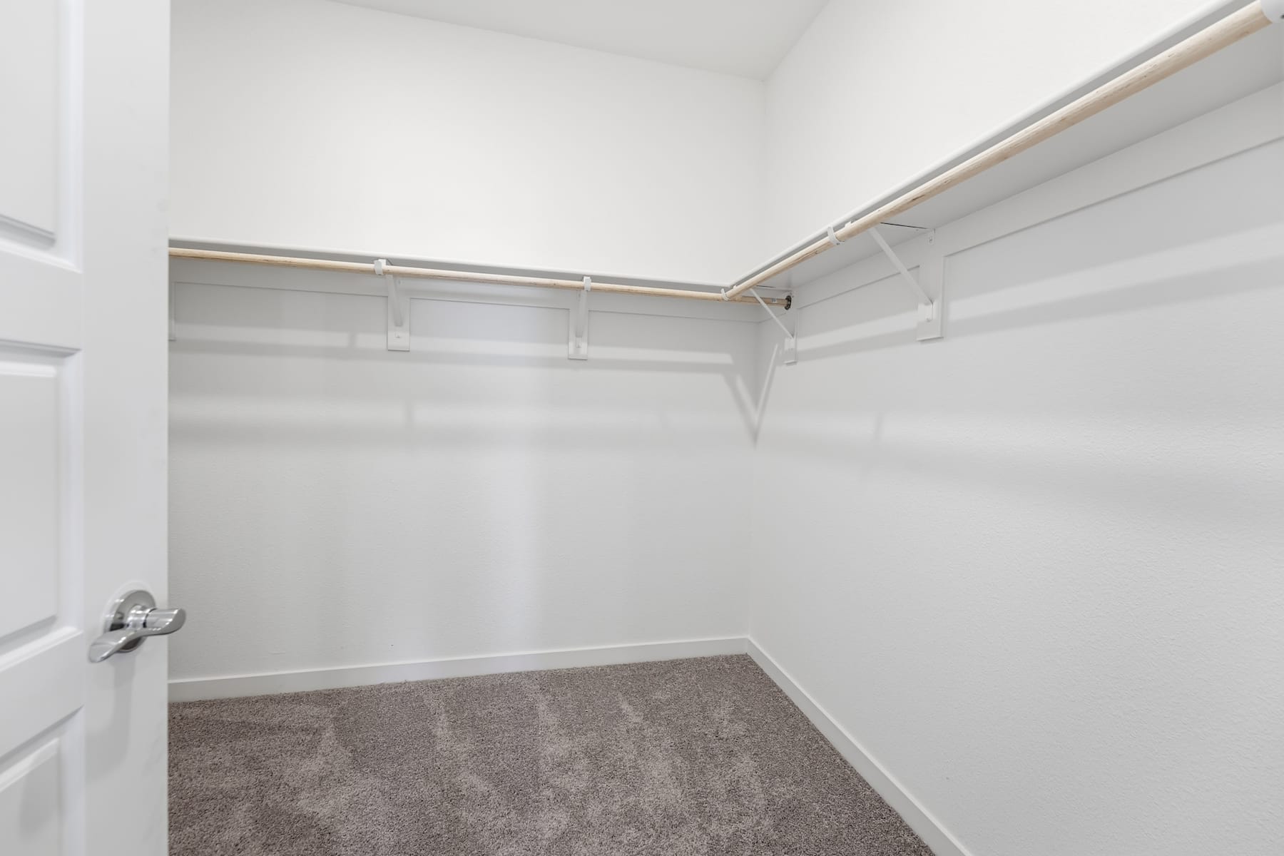 A spacious, well-lit closet with white walls, a wooden floor, and a metal clothing rod running along the top.